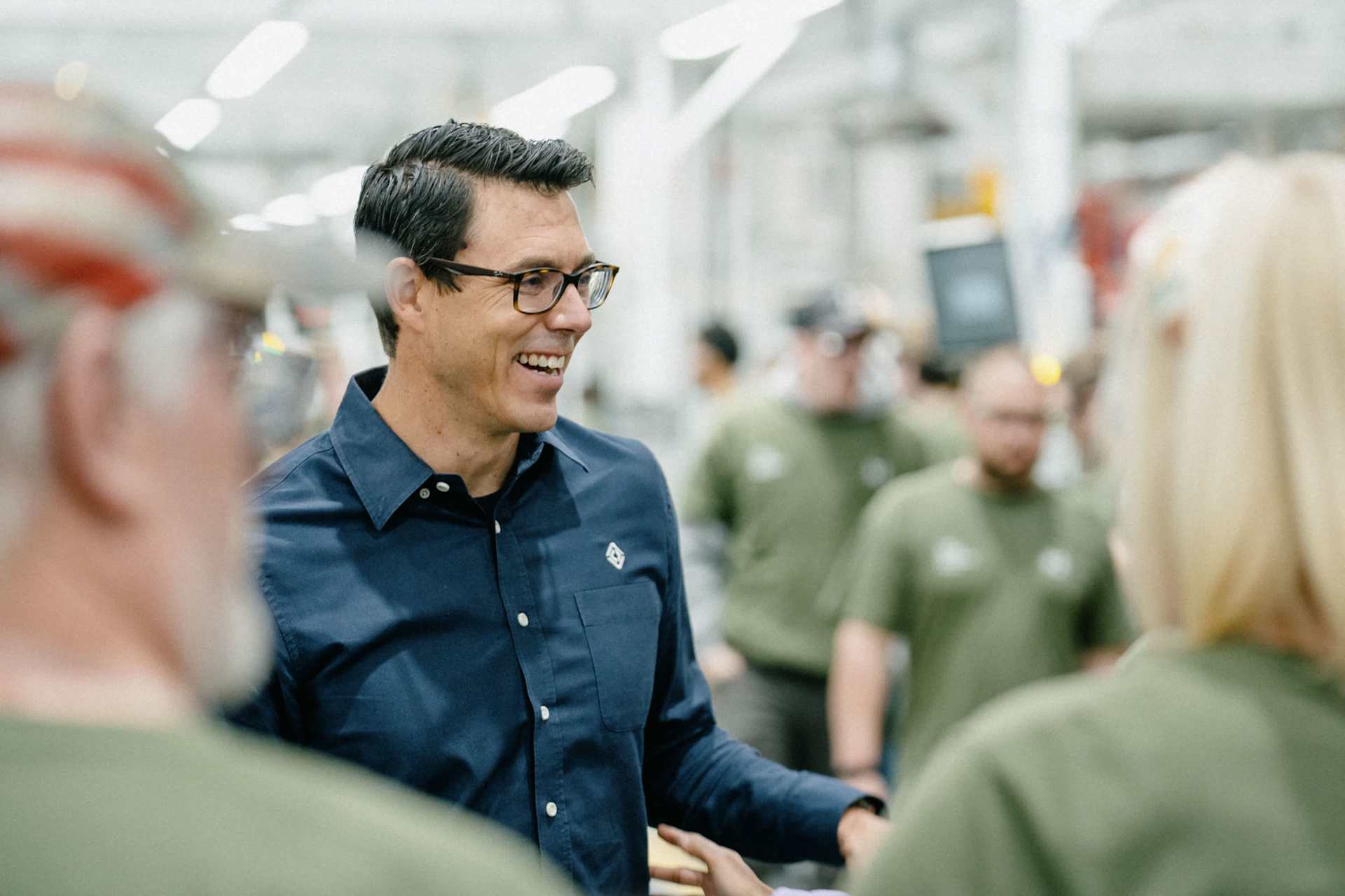 A smiling man in glasses and a Rivian shirt stands in a factory, surrounded by employees in green shirts. The man appears to be addressing the employe