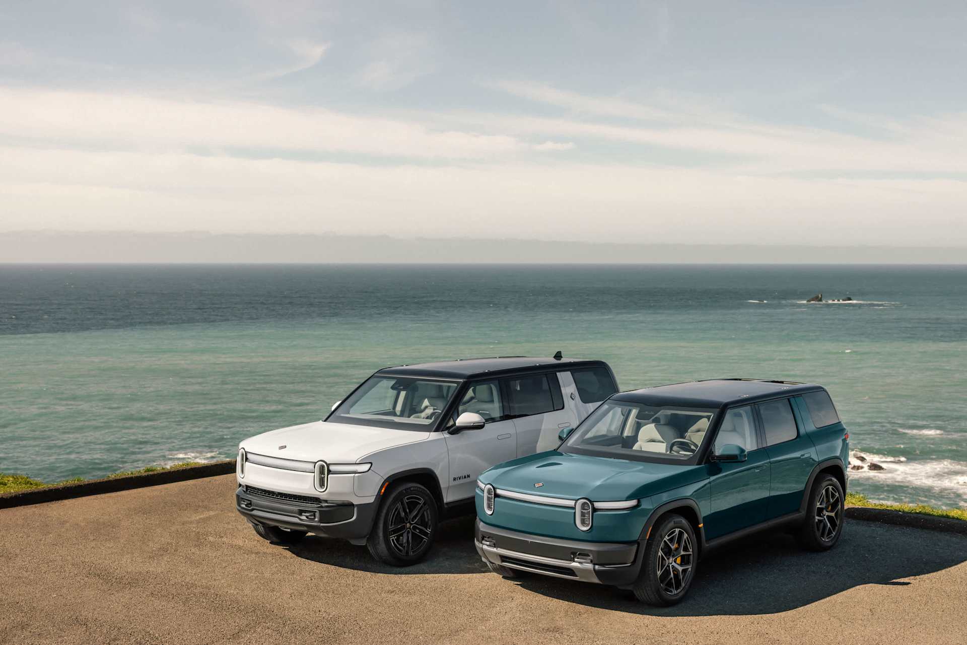 Two Rivian R2 SUVs, one white and one teal, sit parked on a cliffside overlooking the ocean. The white SUV is positioned slightly behind the teal one,