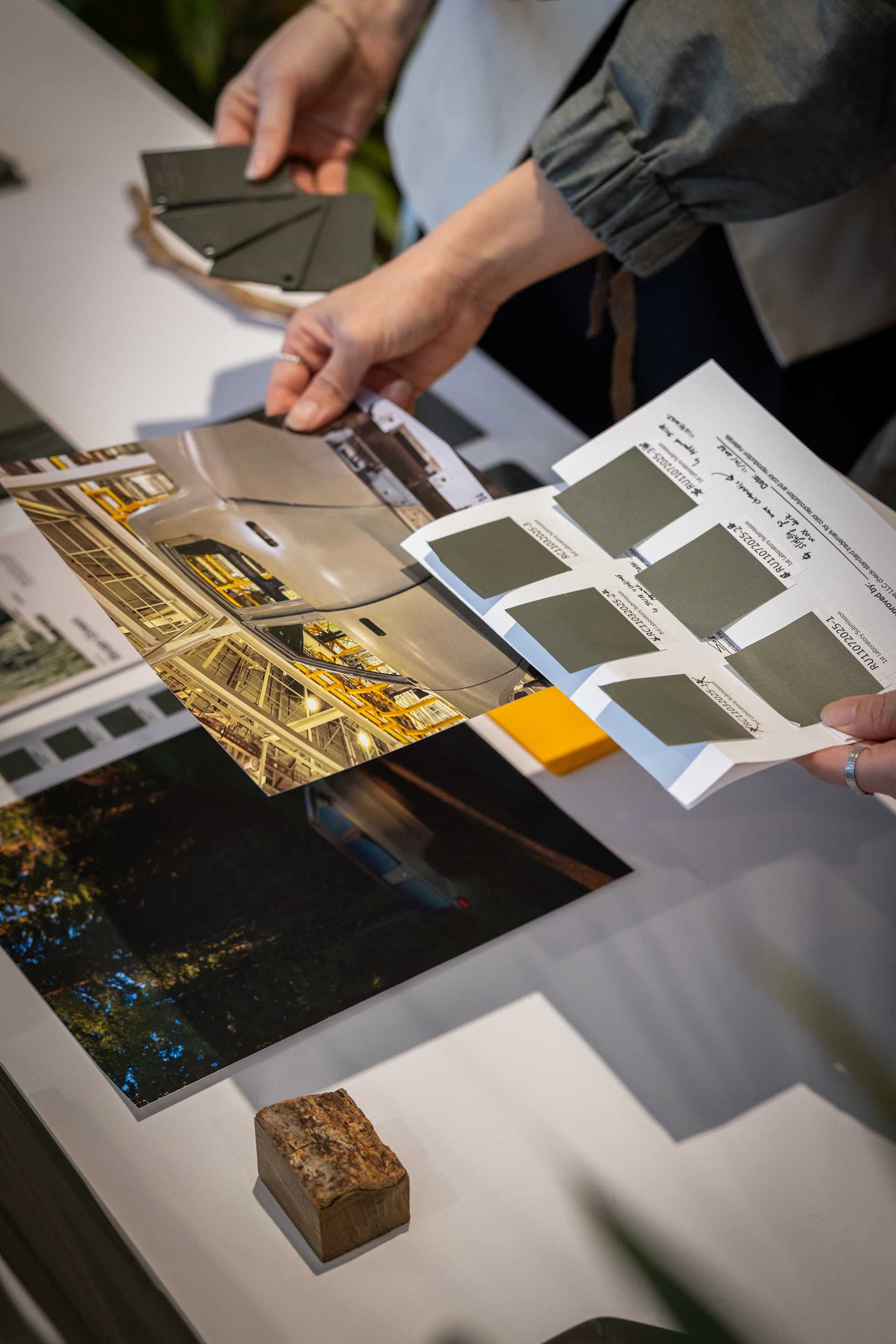 Hands hold color swatches and photos on a white table, comparing tones. One photo shows a car interior, another a nighttime scene. The swatches are ol