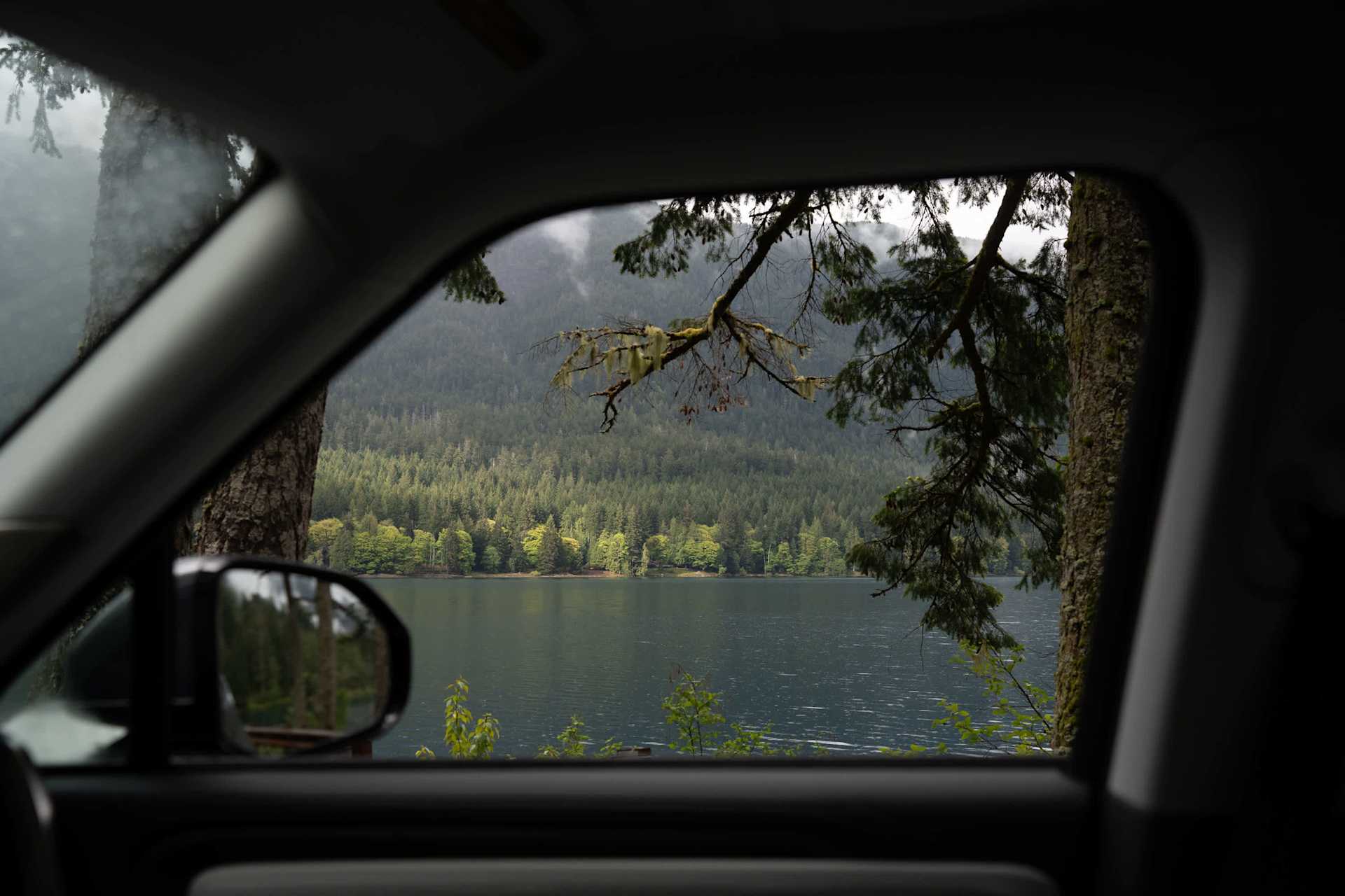 Lake framed by car window and trees. Evergreen forest rises behind still, dark water under cloudy sky. Moss hangs from tree branches.