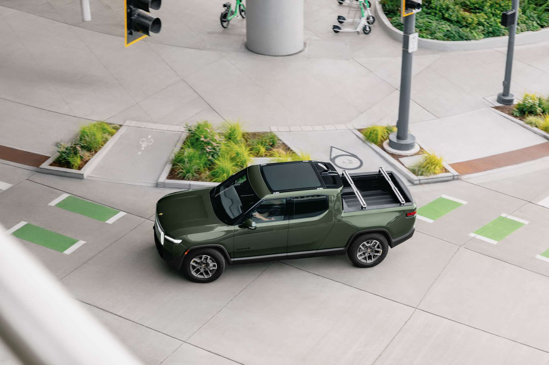 An overhead shot shows a green Rivian truck driving on a city street. The truck approaches a crosswalk with green markings. Scooters are parked near a