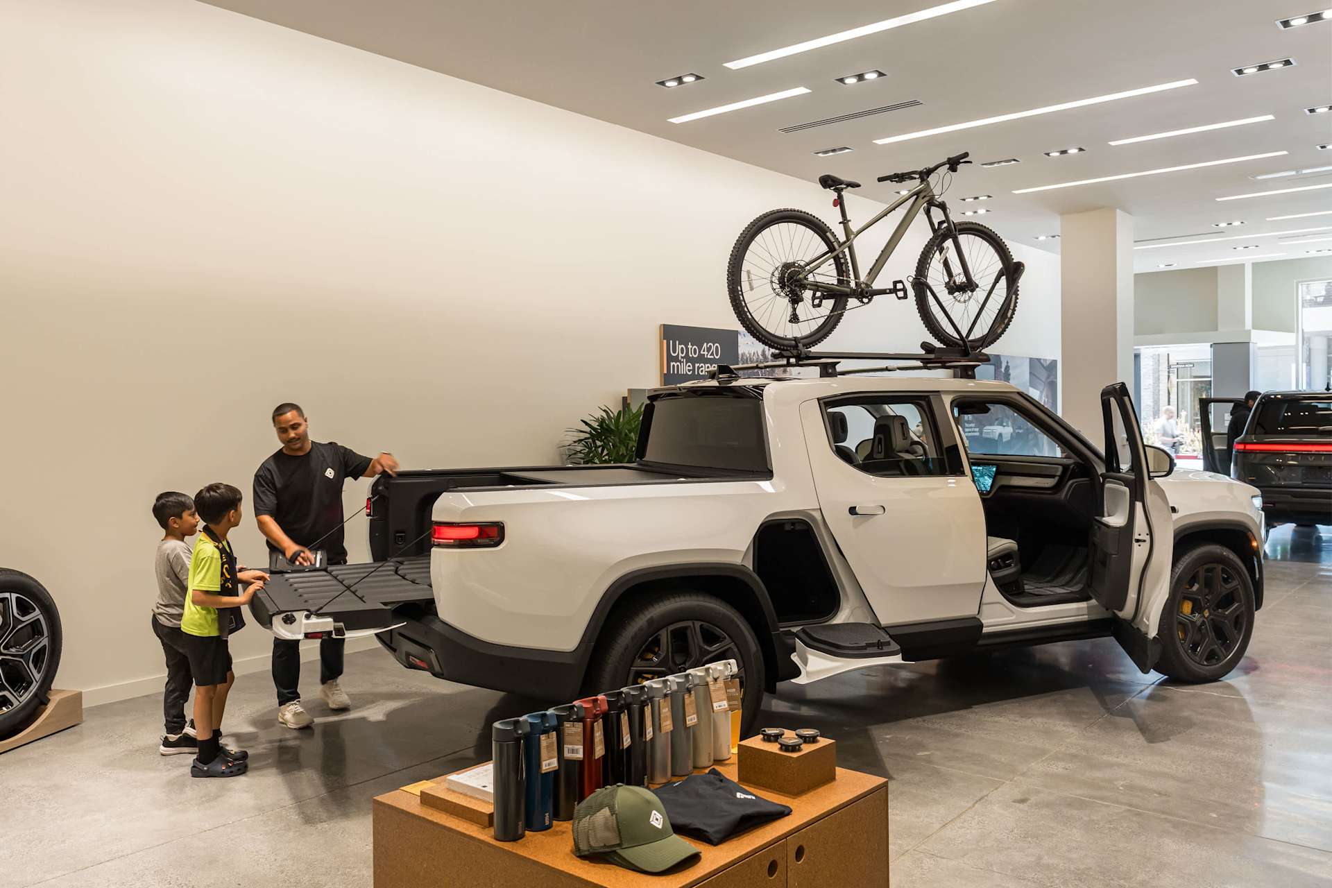 A Rivian employee shows two young boys the extended tailgate of a white R1T pickup truck in a showroom. A mountain bike is mounted on the truck's roof