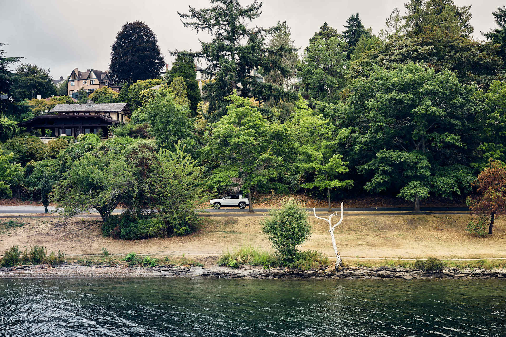 White Rivian R1 drives along a road lined with trees and houses, viewed from across the water. The shoreline is rocky with a grassy bank. Overcast sky