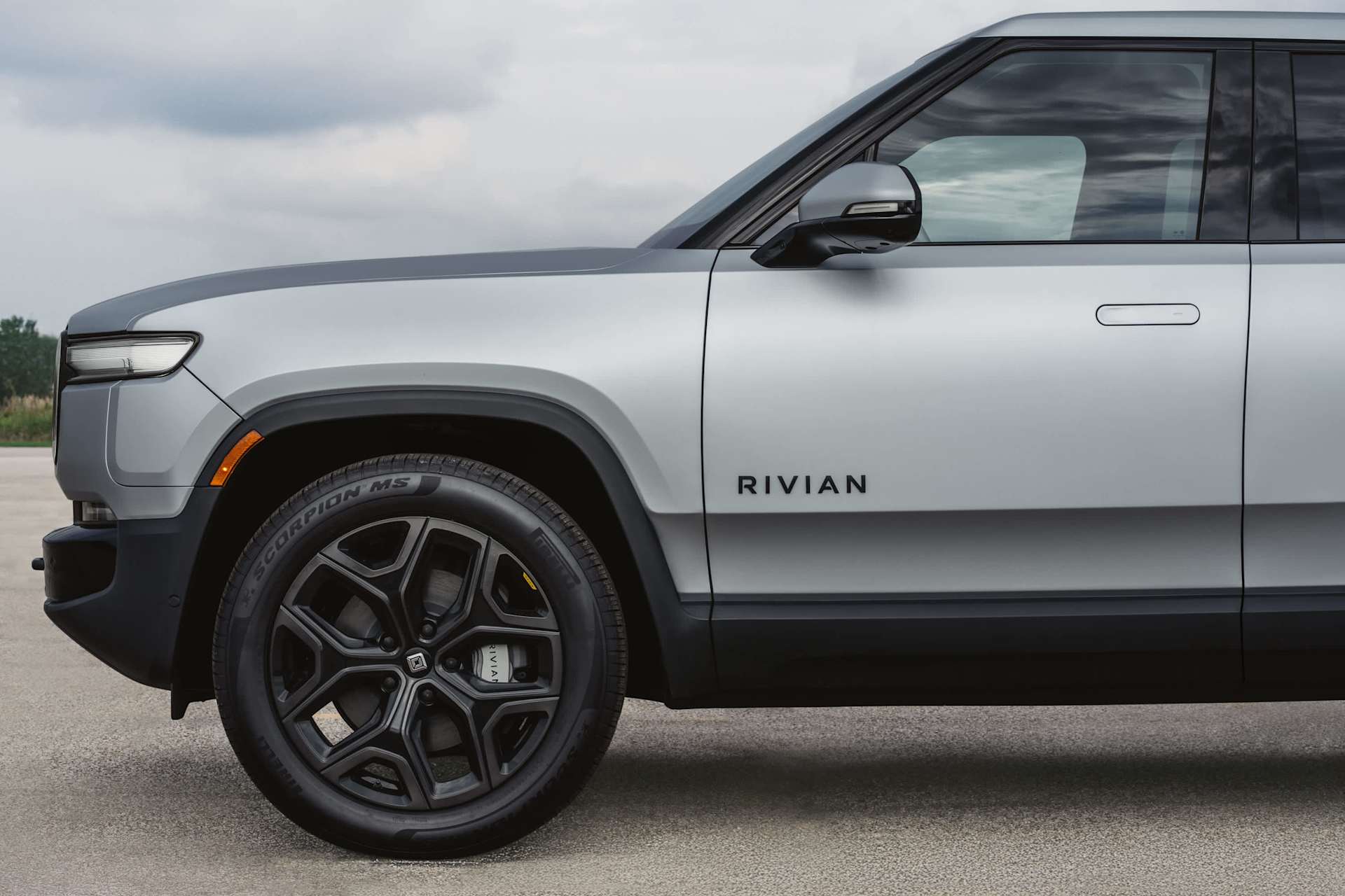 Side view of a silver Rivian vehicle. The Rivian logo is visible on the side panel. The vehicle has black rims and black trim along the bottom.