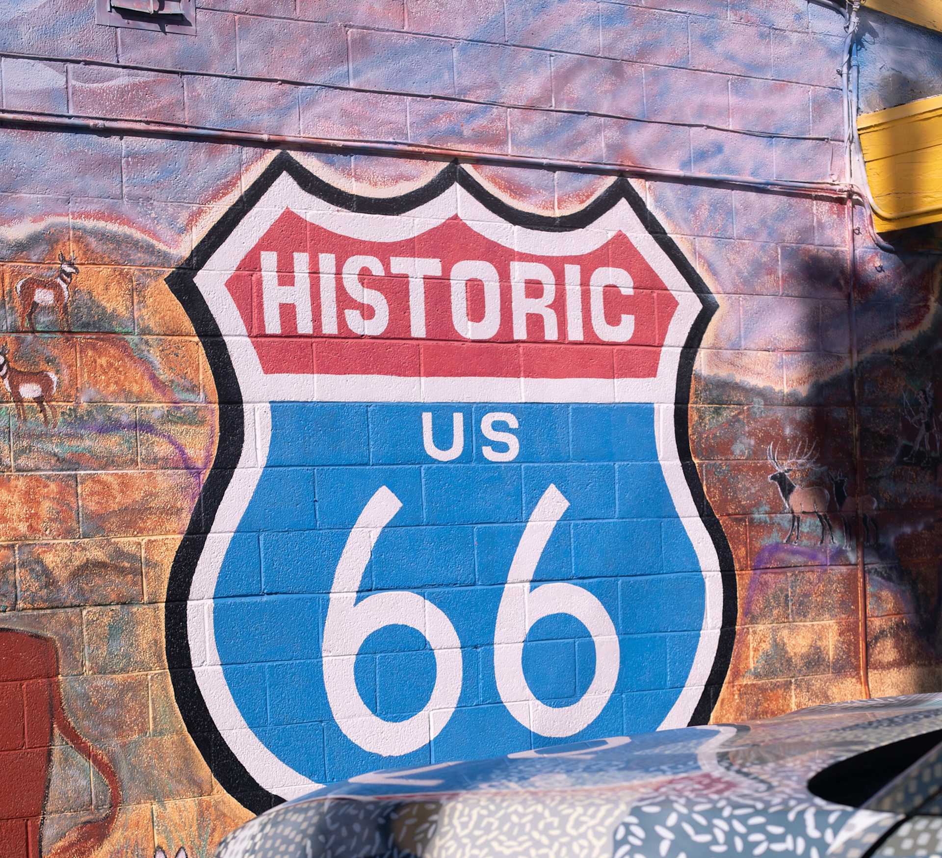 Mural of a Route 66 sign on a brick wall with painted desert landscape and deer. The sign reads "Historic US 66" in red, white, and blue.