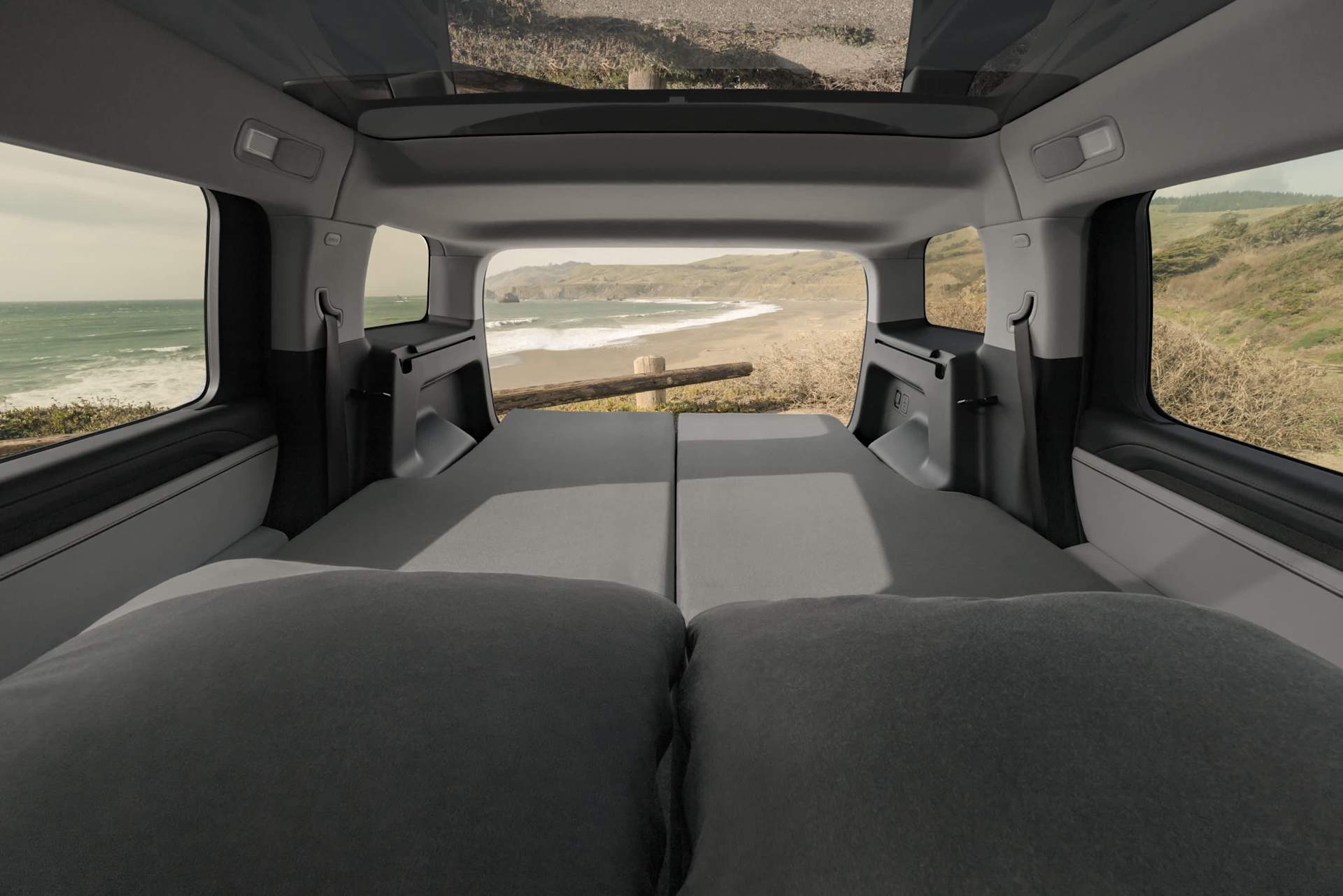 Interior view of a van converted into a camper, with a gray mattress and pillows. The open back doors frame a view of a beach with waves and cliffs.