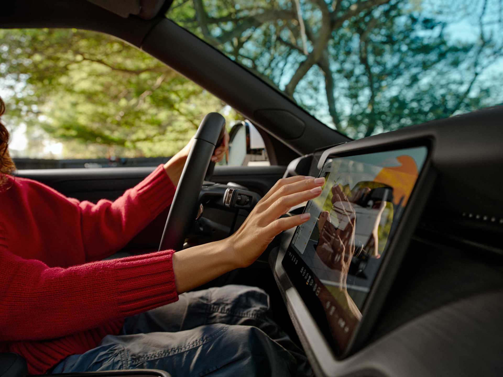 Woman drives a car, touching a large touchscreen display. Trees visible through the windshield suggest daytime driving. She wears a red sweater and gr