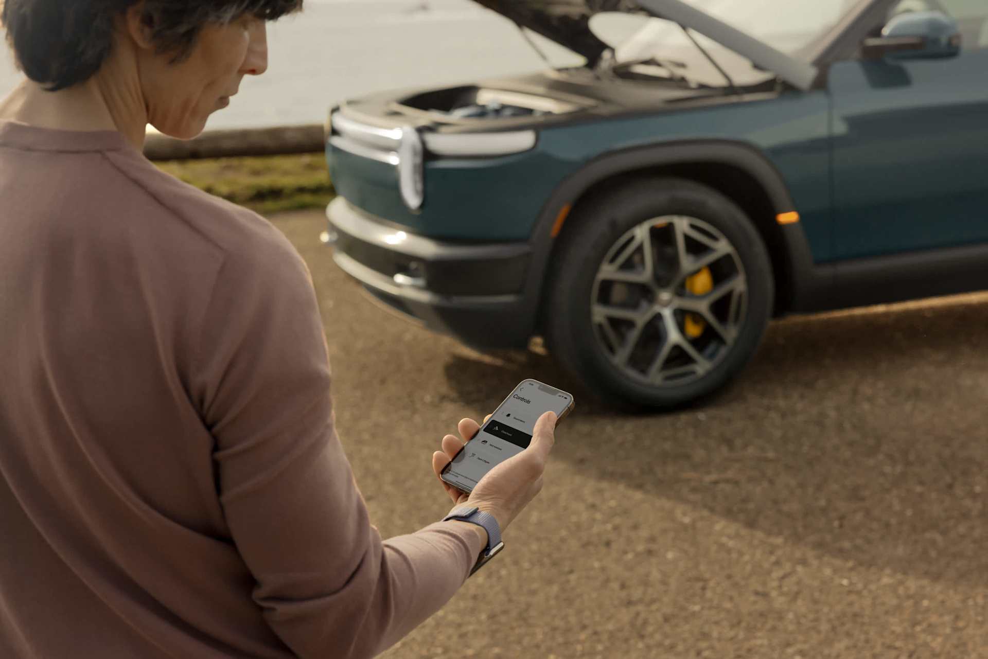 Person uses a smartphone app to control a dark green Rivian truck with its hood open. The setting is outdoors on a sunny day.