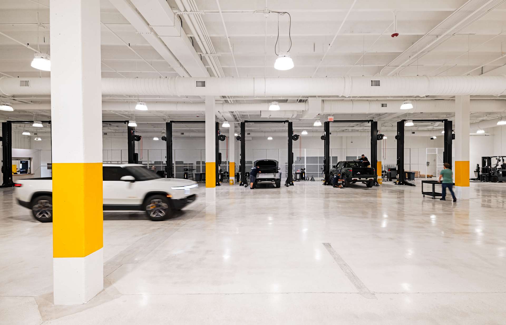 A white Rivian truck drives through a brightly lit service center with several service bays. Technicians work on other Rivian vehicles on lifts in the