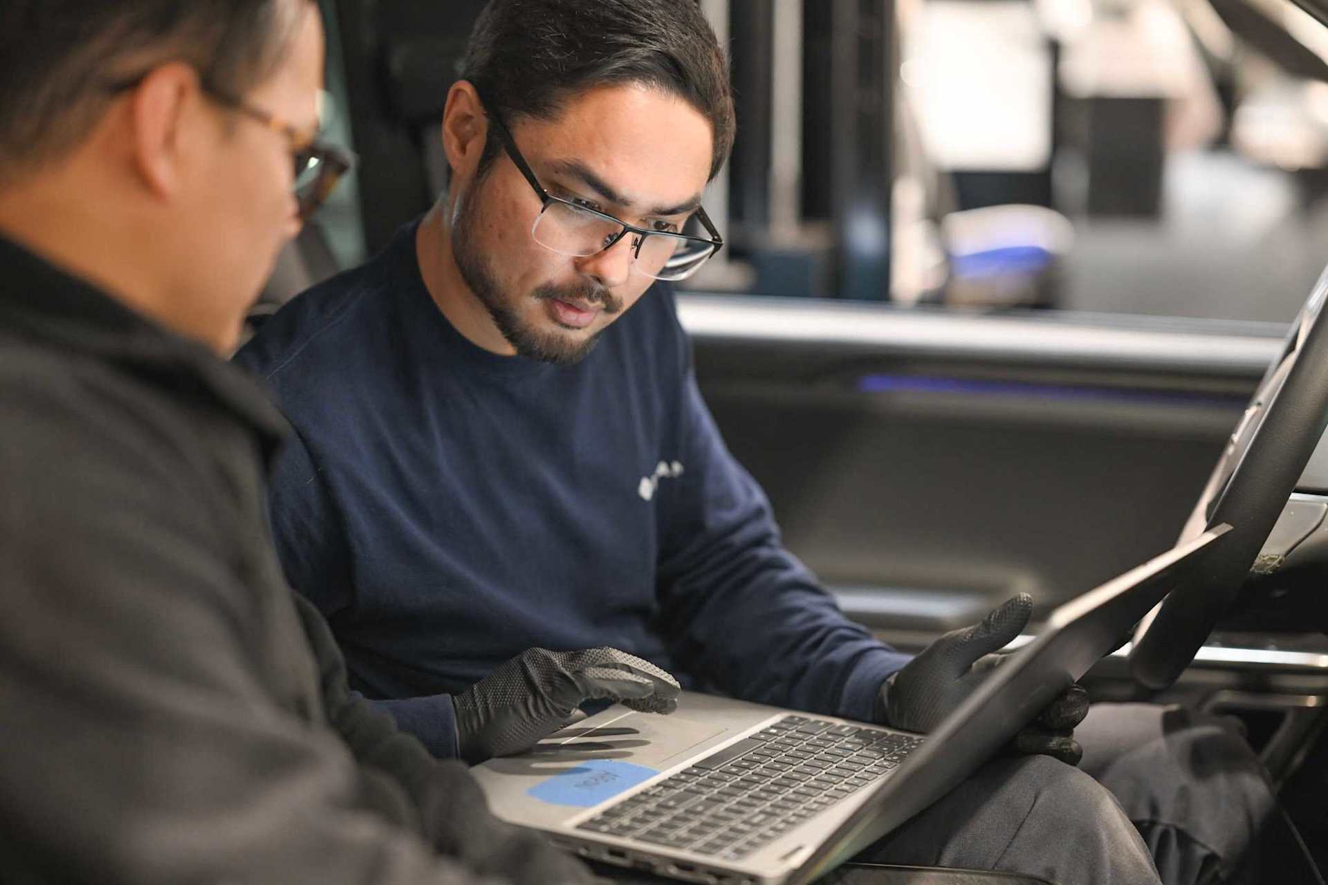 Two Rivian service technicians work inside a vehicle, one types on a laptop. The technician wears black gloves and a navy long-sleeve shirt with the R