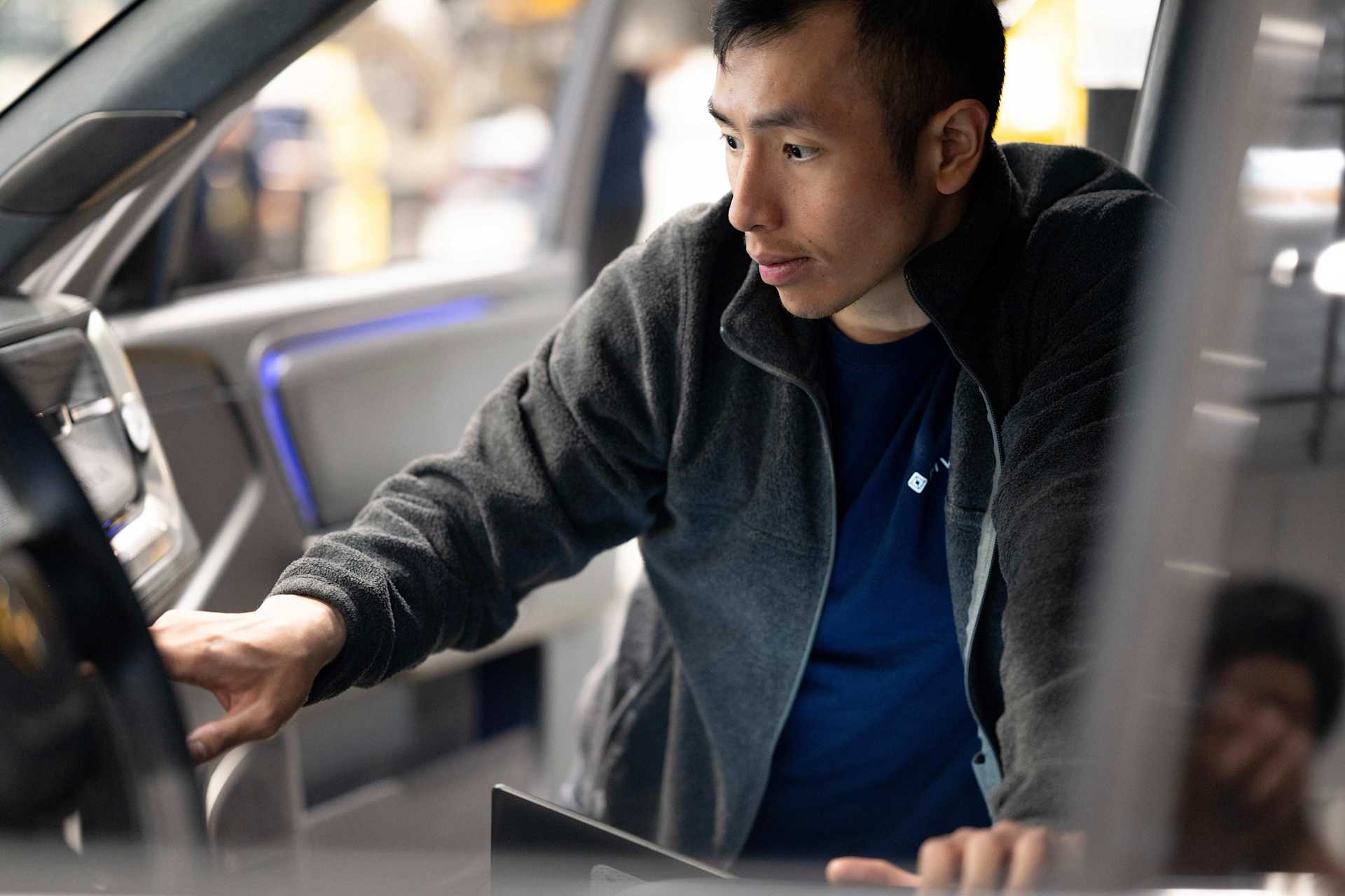 Man inspects the interior of a Rivian vehicle. He wears a blue t-shirt and gray fleece jacket. Interior lights illuminate the dashboard.