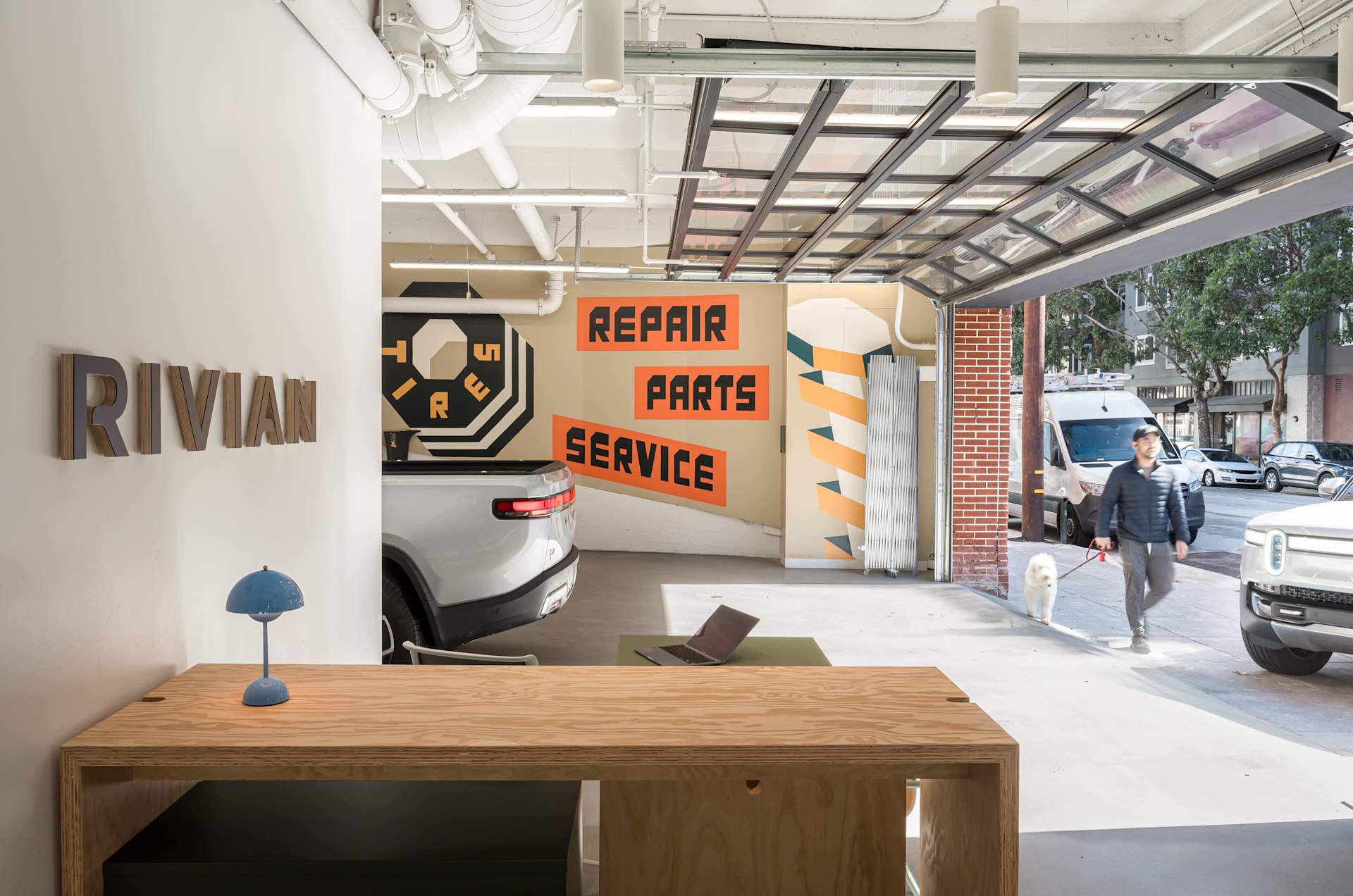 Rivian service center with open garage door. Man walks dog outside. Interior wall reads "Repair, Parts, Service" in orange block letters. A Rivian tru