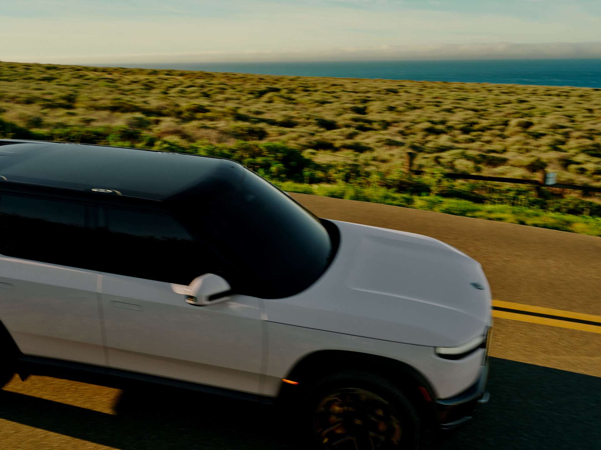 White Rivian SUV drives on a paved road. The vehicle has a dark roof and is traveling past a grassy hillside overlooking the ocean under a blue sky.