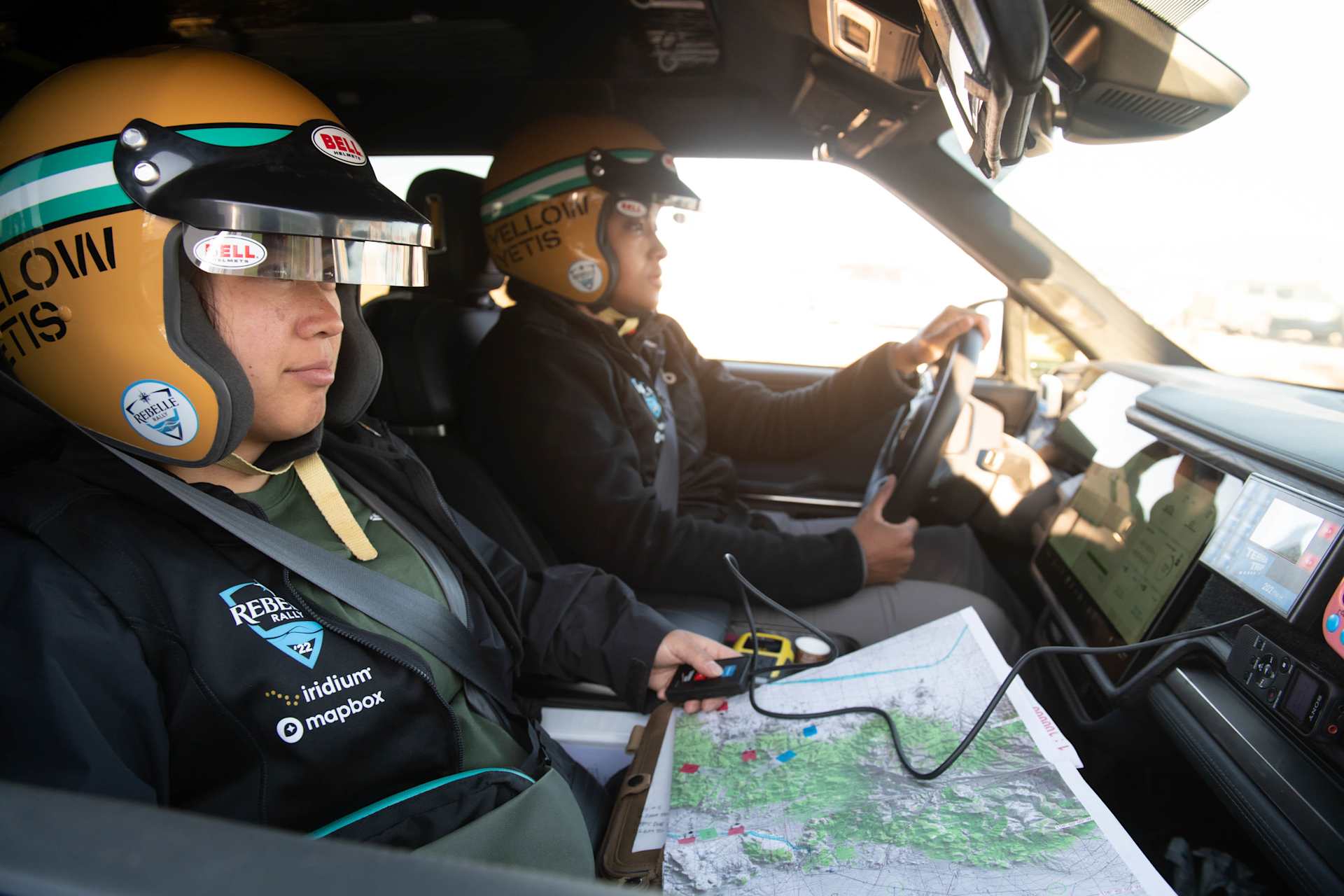 Inside a Rivian, two people wearing "Yellow Yetis" helmets navigate with a map and handheld device. The driver grips the steering wheel, while the nav