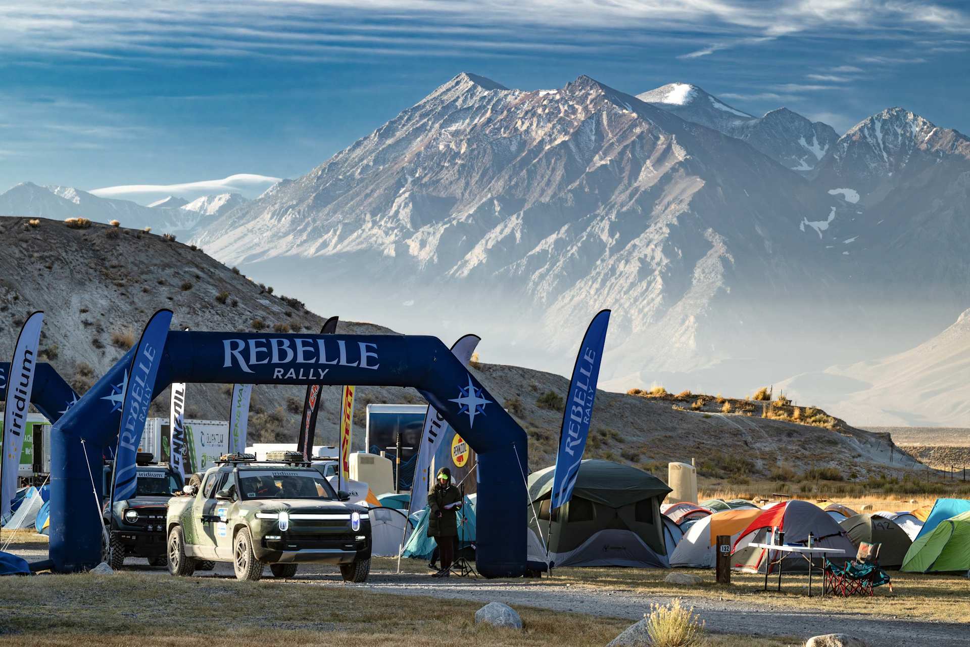 Rivian truck drives under "Rebelle Rally" arch at a desert basecamp with tents, mountains in the background.
