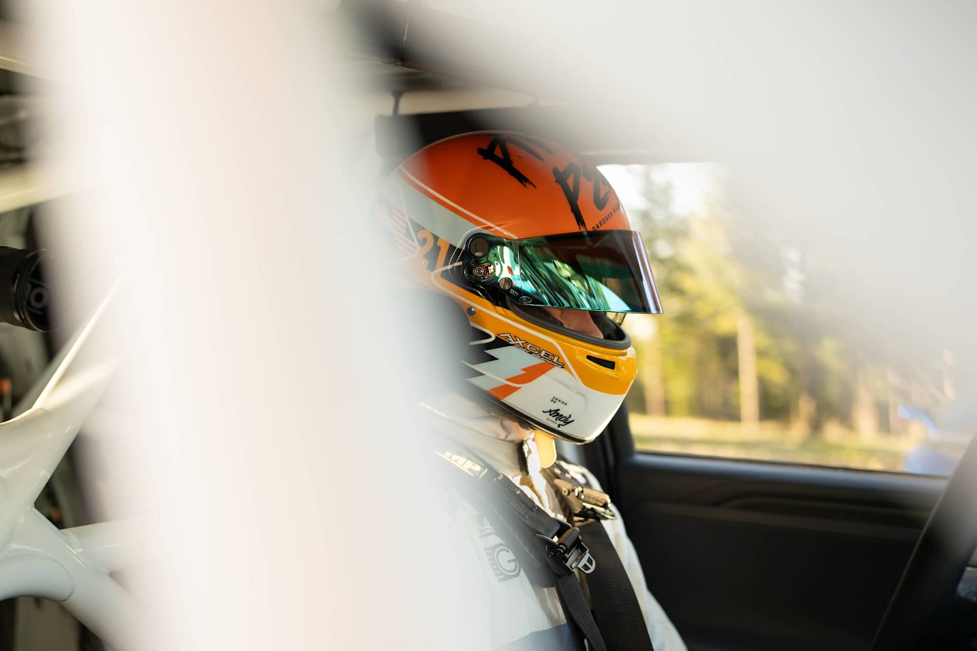 Race car driver sits in vehicle wearing orange helmet with visor, harness, and white racing suit. Trees are visible through the car window.