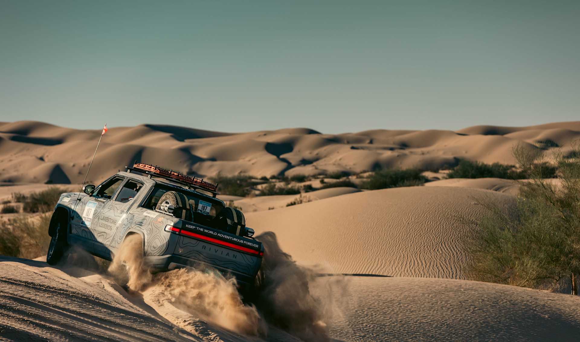 Rivian truck ascends a sand dune, kicking up sand. The truck has a topographical map design and roof rack with spare tires. Desert landscape, clear sk