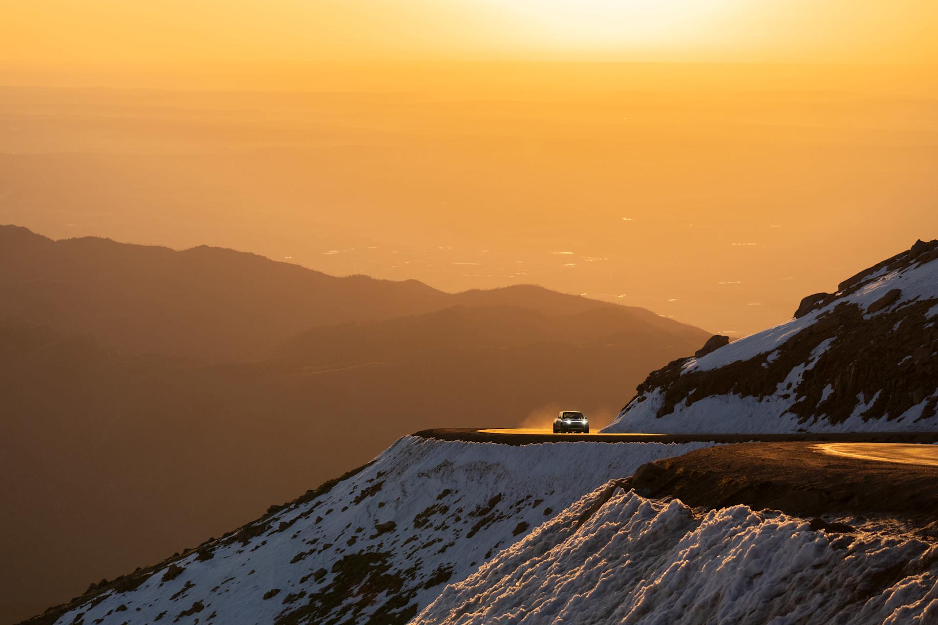 Rivian drives along a mountain road at sunset. The road is lined with snow and the sky is a warm orange.