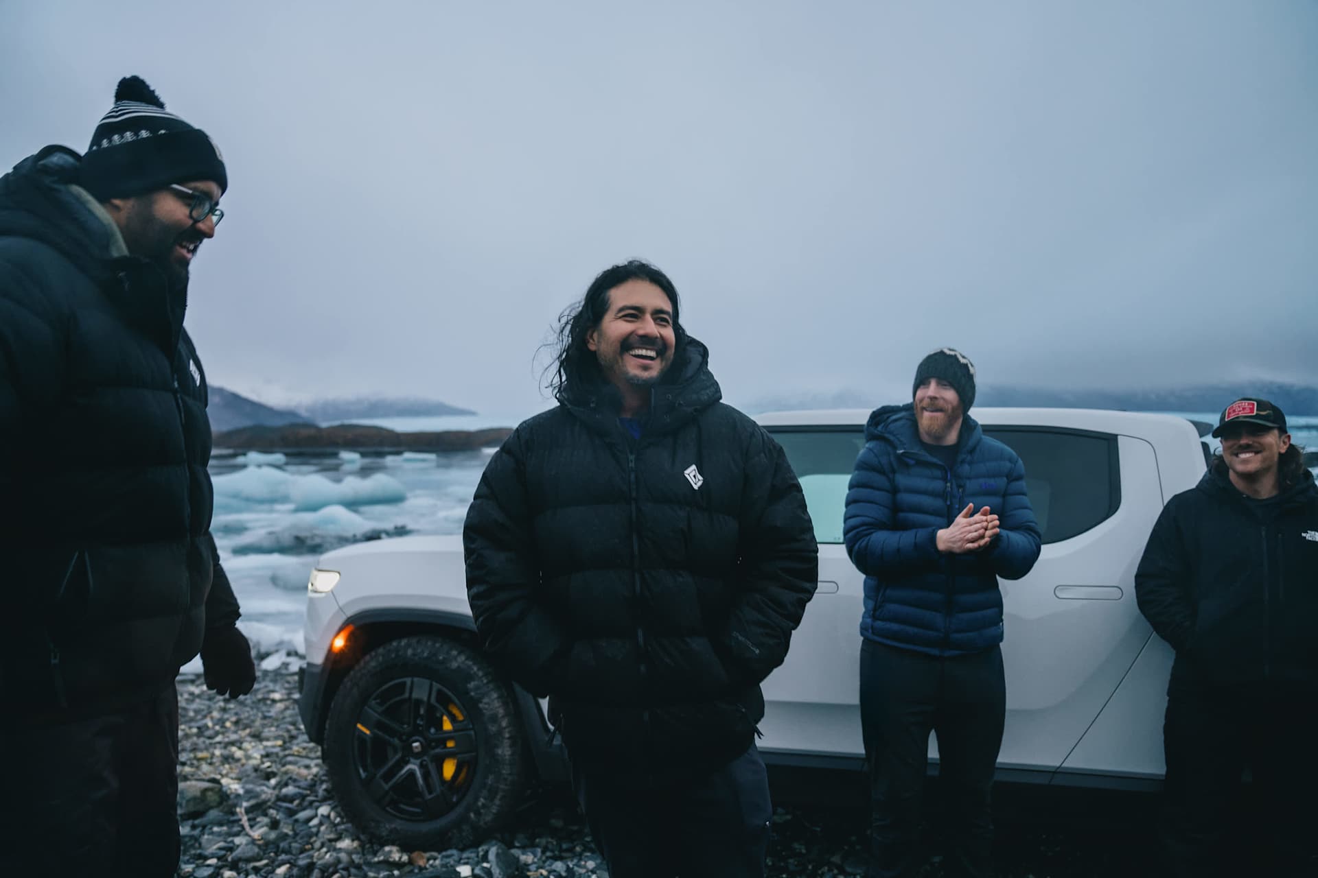 Four smiling people stand near a Rivian truck on a rocky shore with icebergs in the background. They wear winter jackets and hats. Overcast sky.