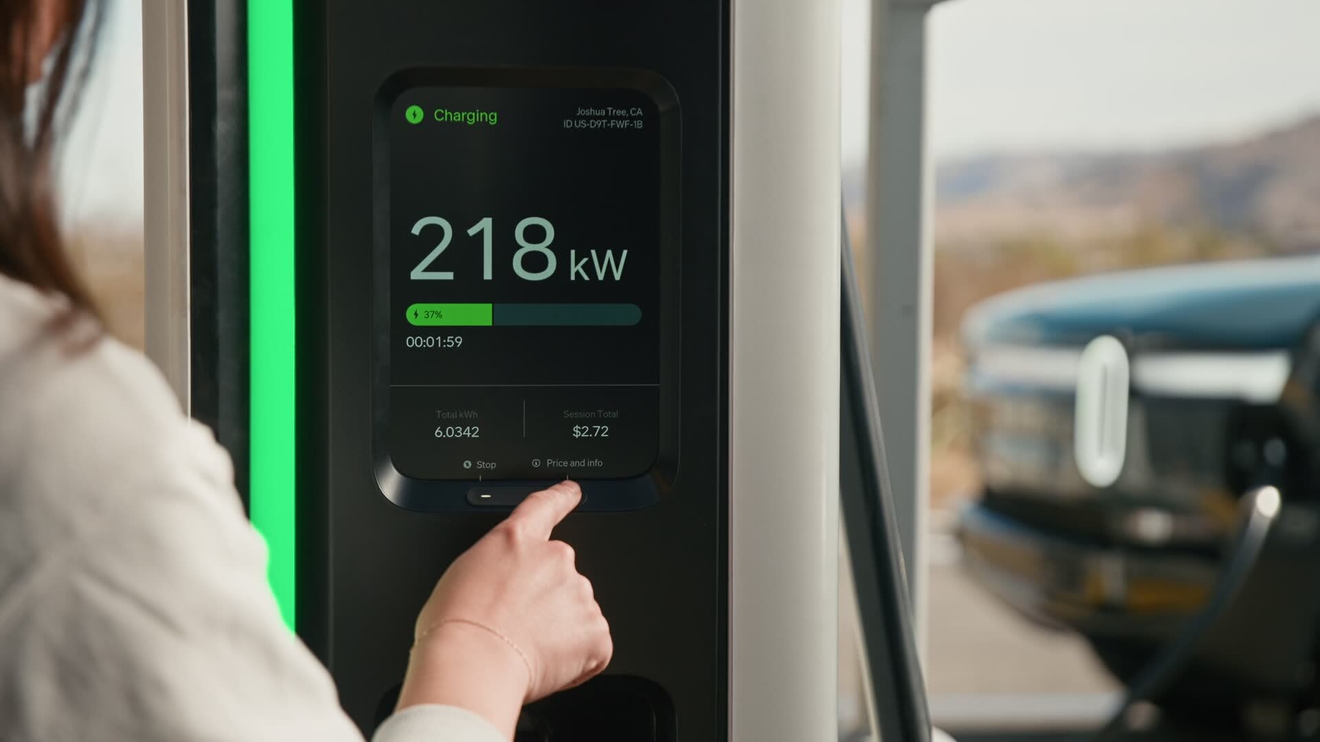 Person touches "Price and info" on an EV charging station screen displaying "Charging" and "218kW" in Joshua Tree, CA. A blurred EV is visible in the 