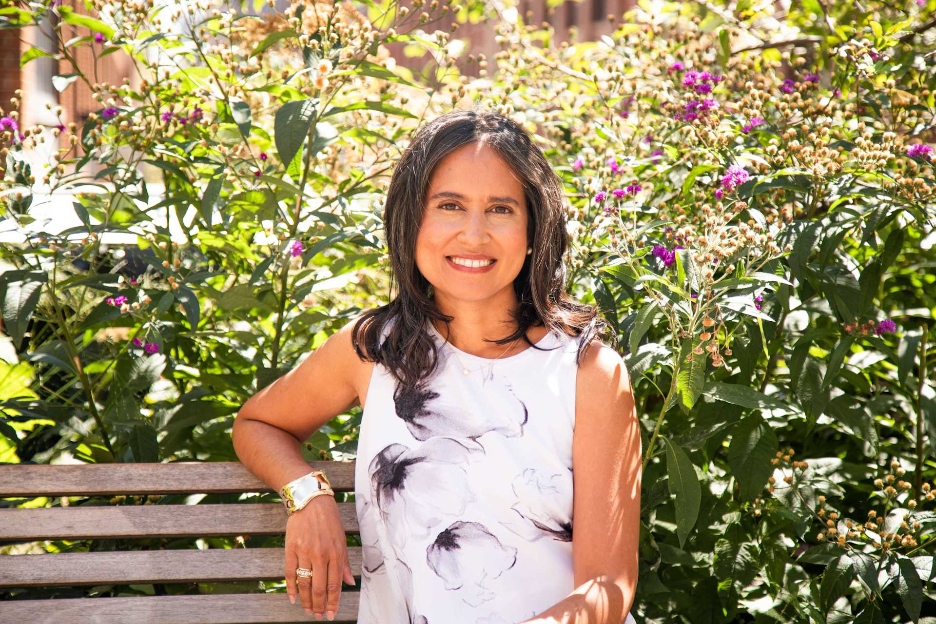 Woman smiles, seated on a wooden bench in front of flowering bushes. She wears a sleeveless white dress with gray floral pattern and gold jewelry. Sun