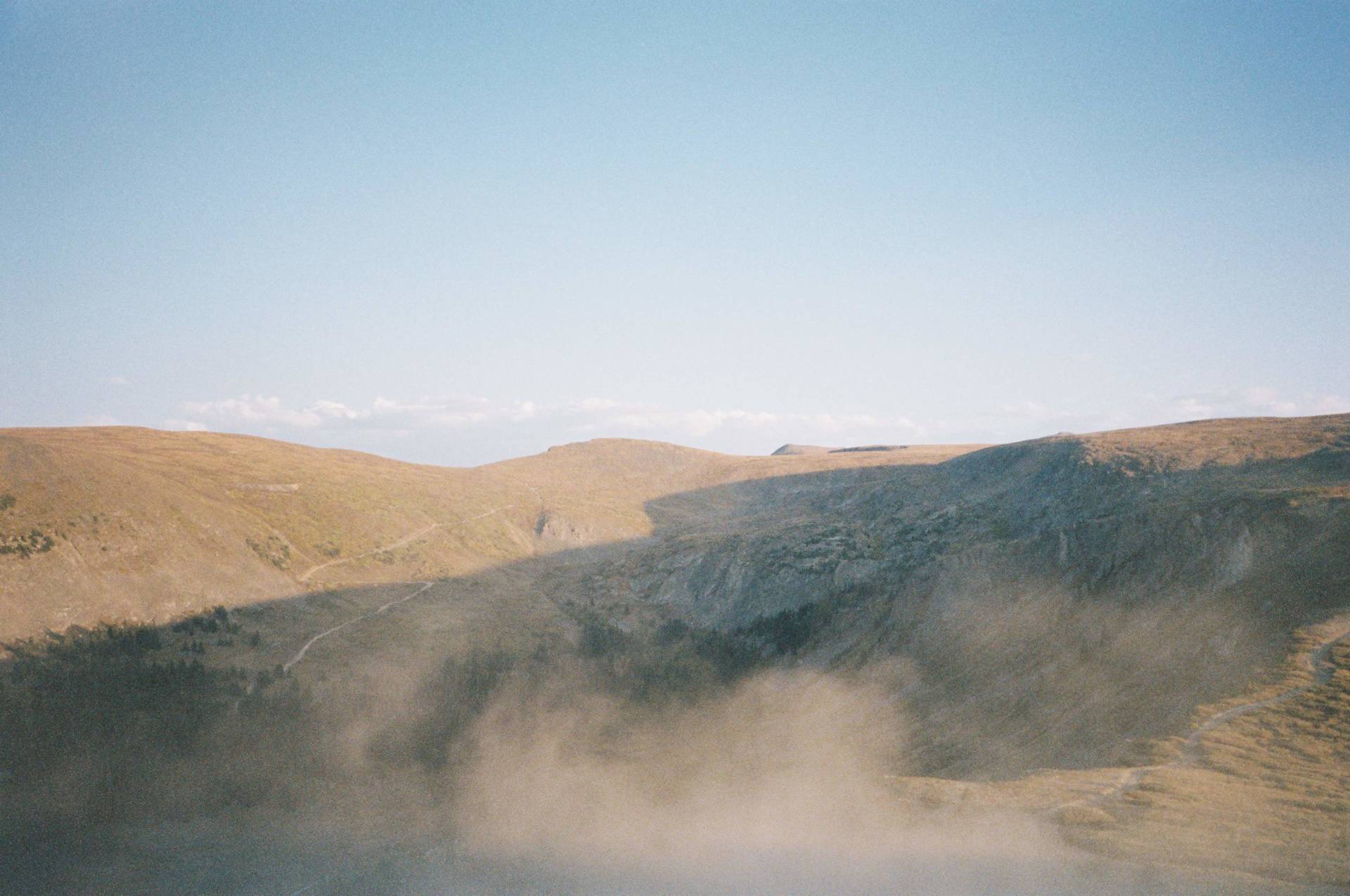 Hillsides with sparse vegetation and a winding path are partially obscured by low-lying fog under a clear sky.