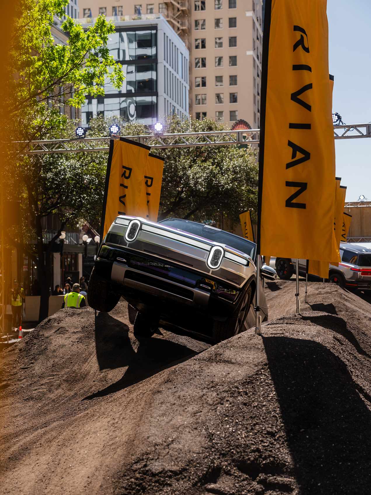 Rivian truck climbs a dirt hill at SXSW 2026. Yellow Rivian flags line the course. City buildings are in the background on a sunny day.