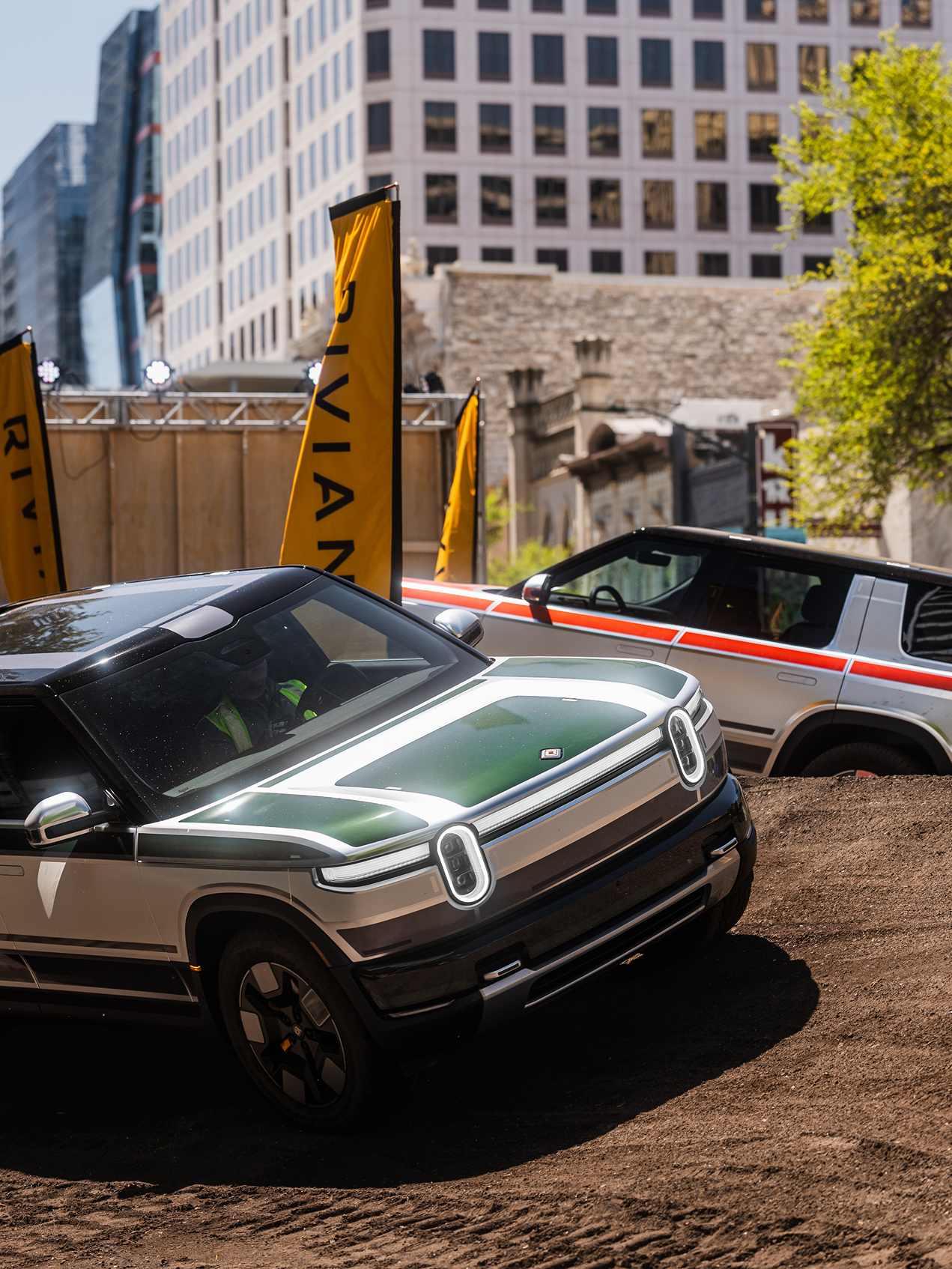 Rivian vehicles on a dirt track at SXSW 2026. The front vehicle is green and white, with yellow Rivian flags and Austin buildings in the background.