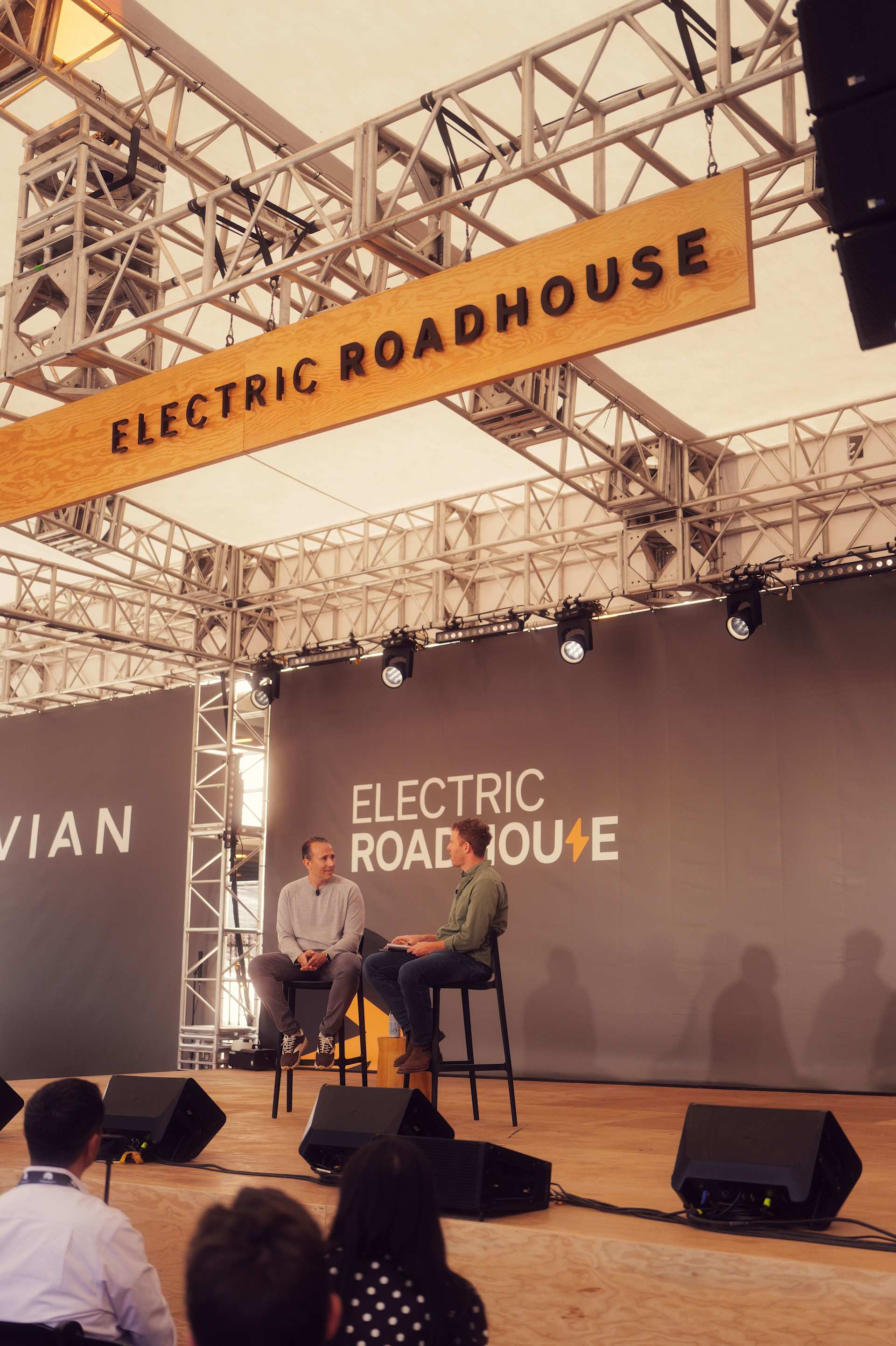Two men sit on stools on a stage at "Electric Roadhouse" during SXSW 2026. The men face each other in conversation. A banner with "Electric Roadhouse"