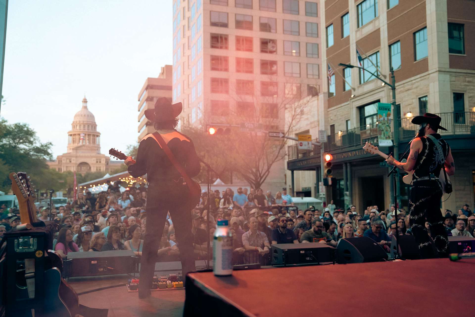 Two musicians perform on an outdoor stage for a large crowd at SXSW 2026. The Texas State Capitol building is visible in the background.
