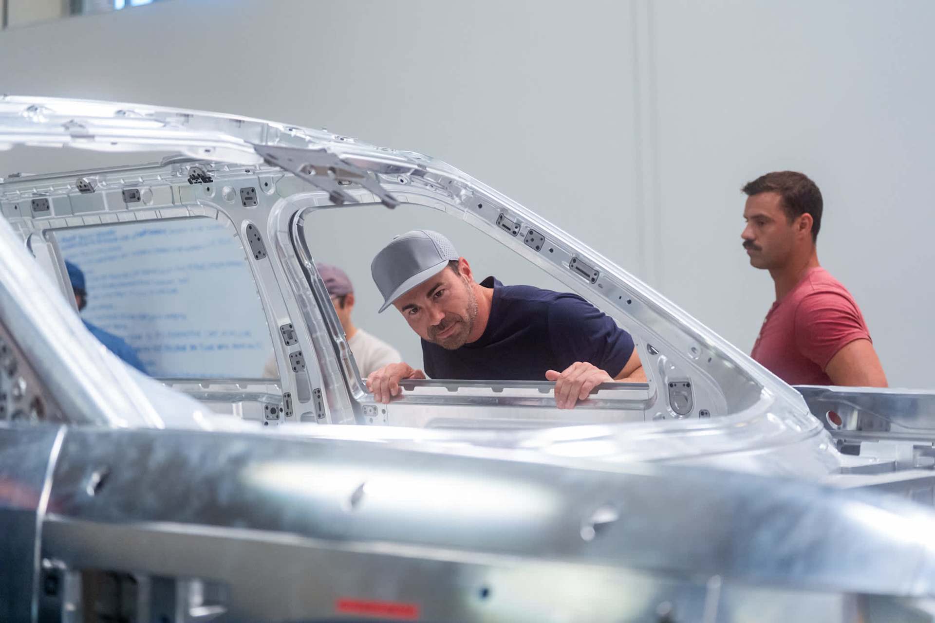 Man in baseball cap peers through the frame of a Rivian vehicle under construction in a bright factory setting, another man stands behind him.