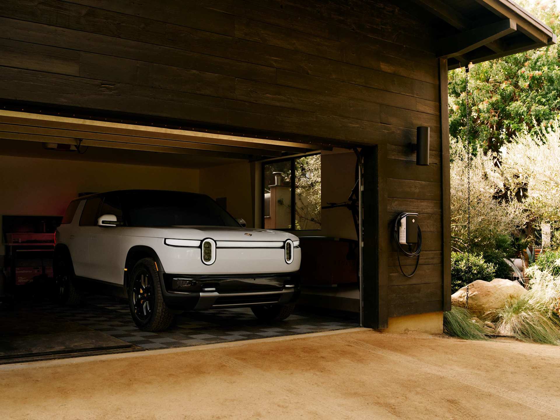 White Rivian SUV parks in a dark wood garage with the door partially open. A home charging unit is mounted on the garage exterior next to green foliag