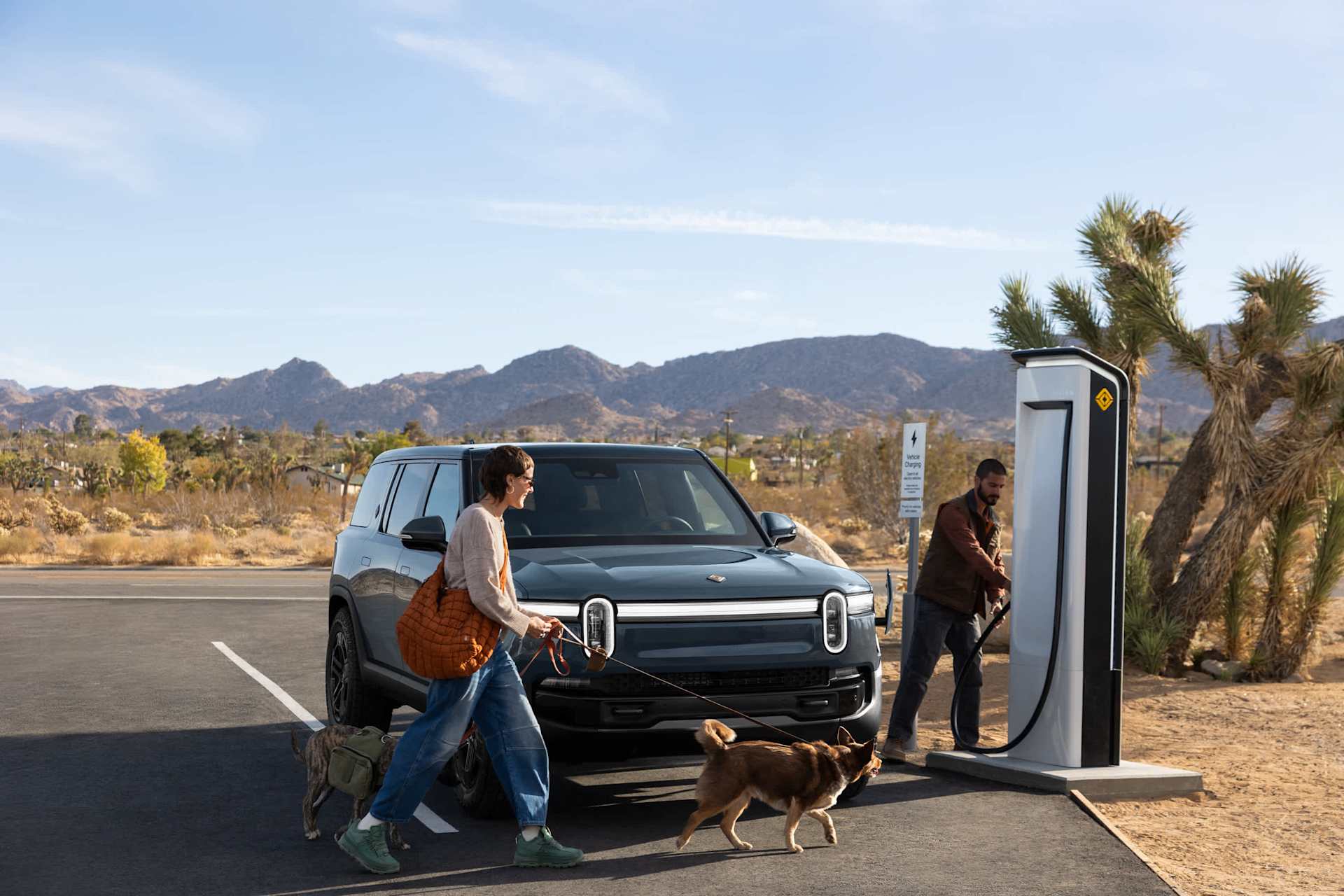 Woman walks two dogs past a Rivian SUV while a man charges it at a charging station. Desert landscape with mountains in the background on a clear day.