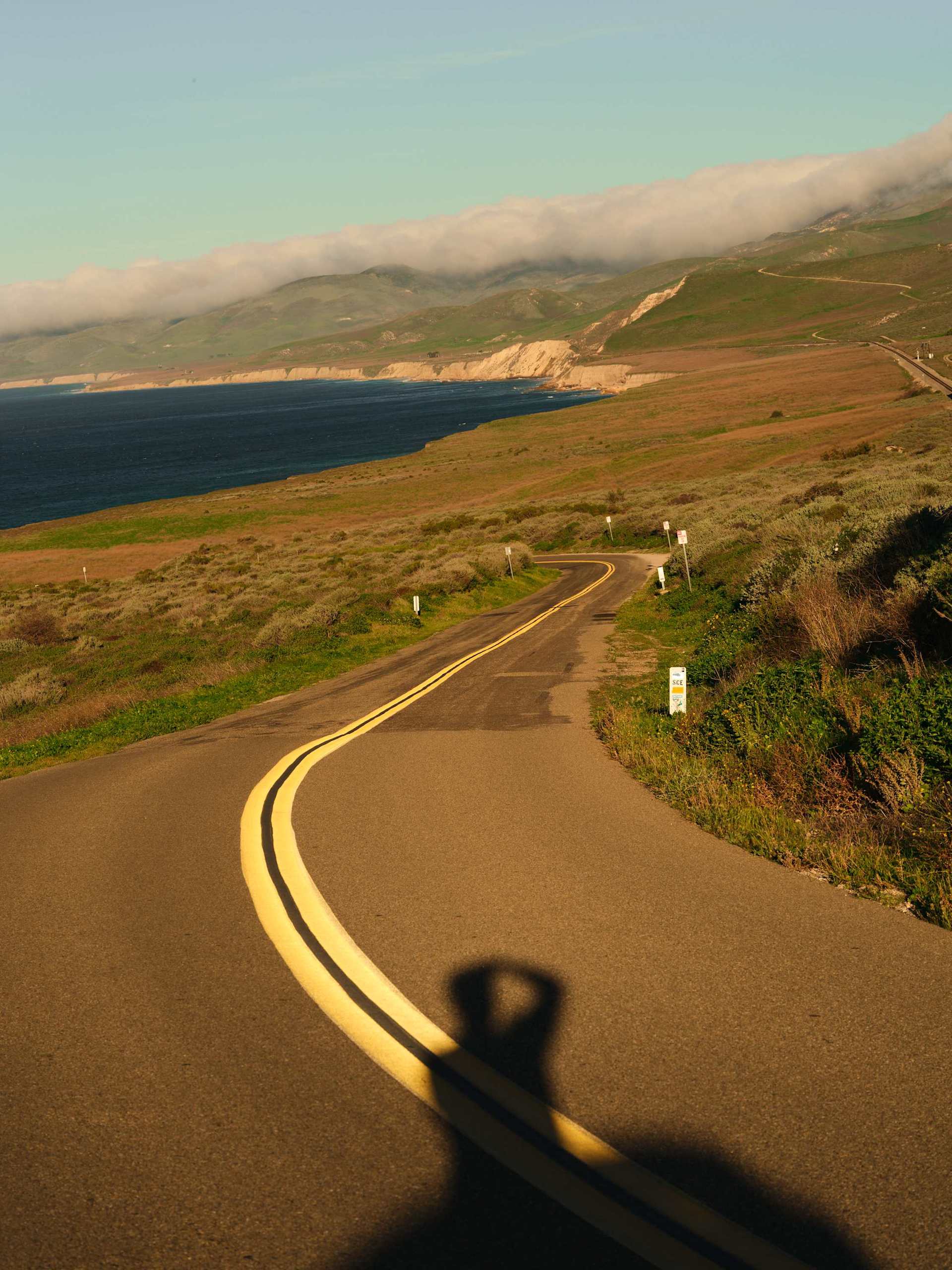 Winding coastal road with double yellow lines curves toward the horizon. The Pacific Ocean meets grassy hills under a cloudy sky. Shadow of a person t