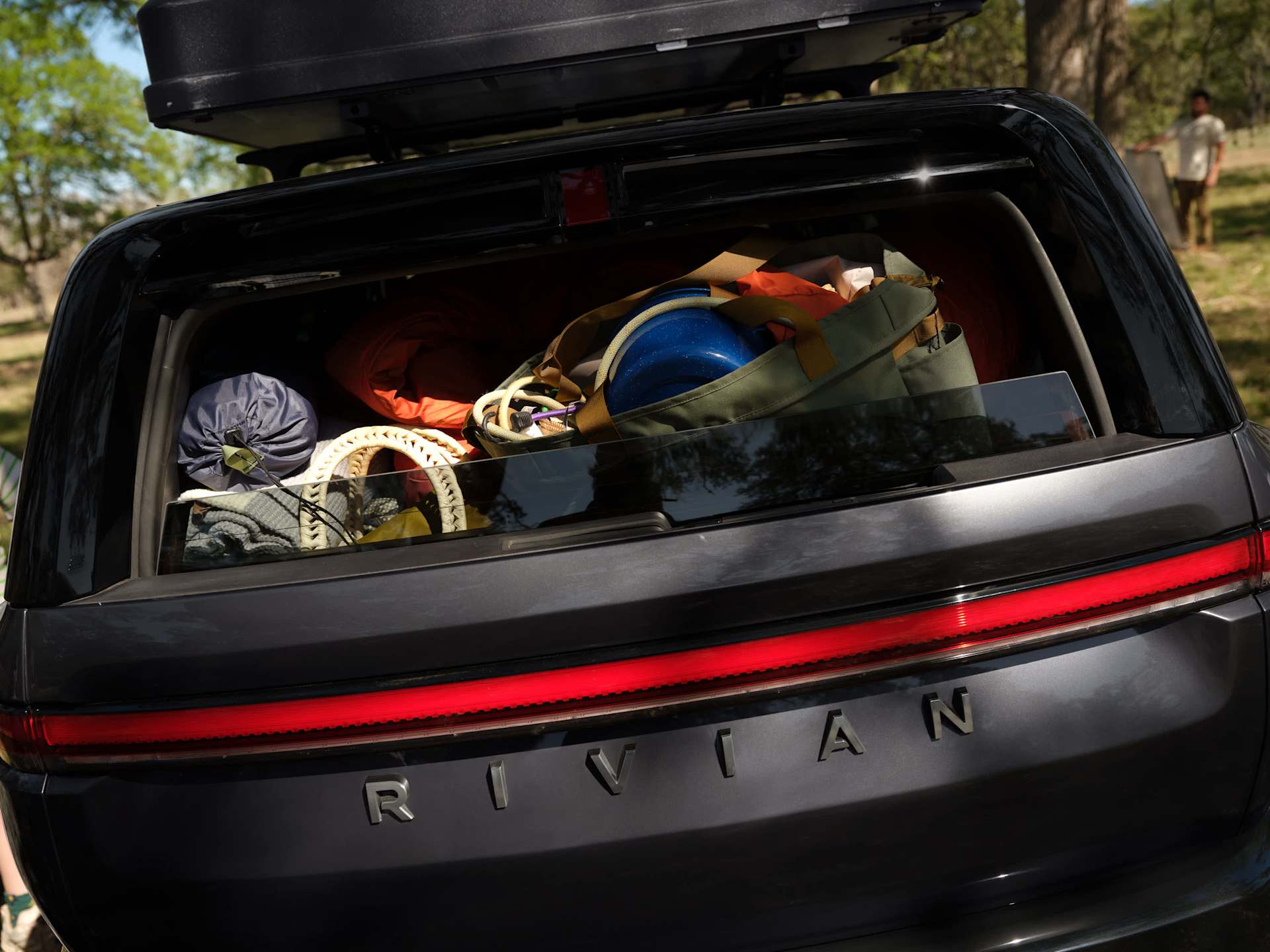 Rear view of a gray Rivian SUV packed with camping gear. The open trunk reveals a tent, sleeping bag, cooking equipment, and climbing ropes. "RIVIAN" 