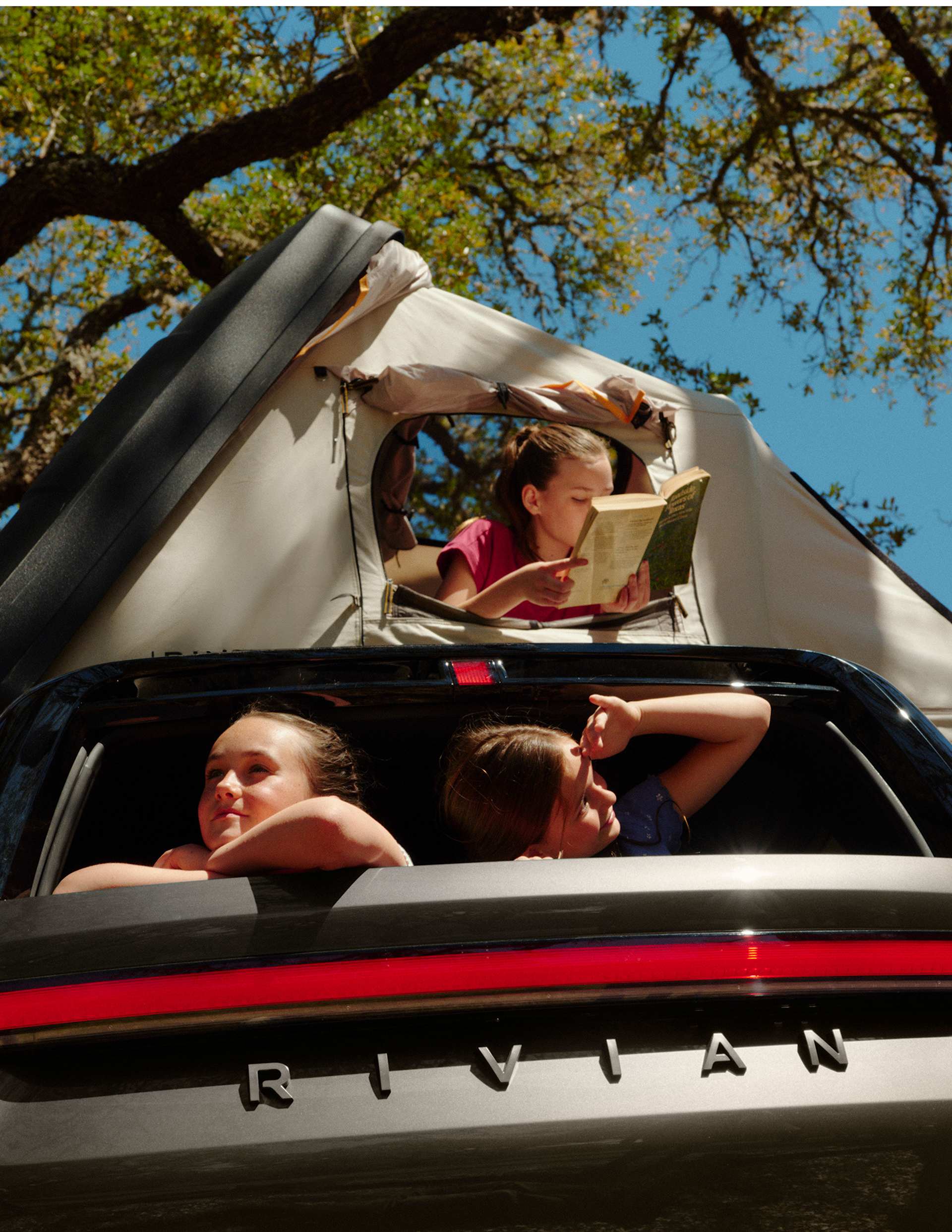 Three girls in a Rivian truck with a tent on top. One girl reads a book inside the tent, while the other two girls look out the back window of the tru