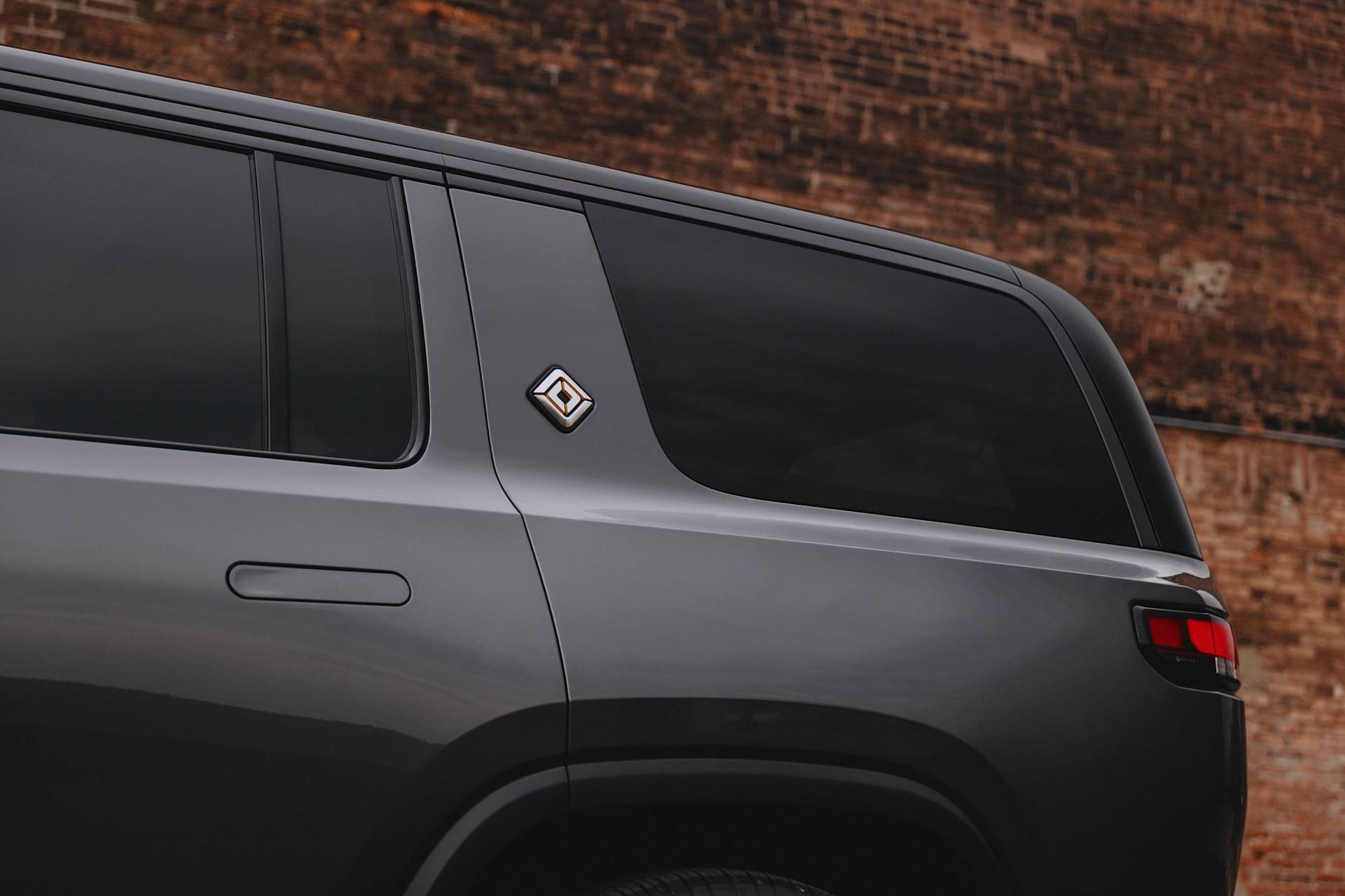 Close-up of a gray Rivian SUV parked against a brick wall. The vehicle's logo, a stylized compass, is visible on the rear pillar.
