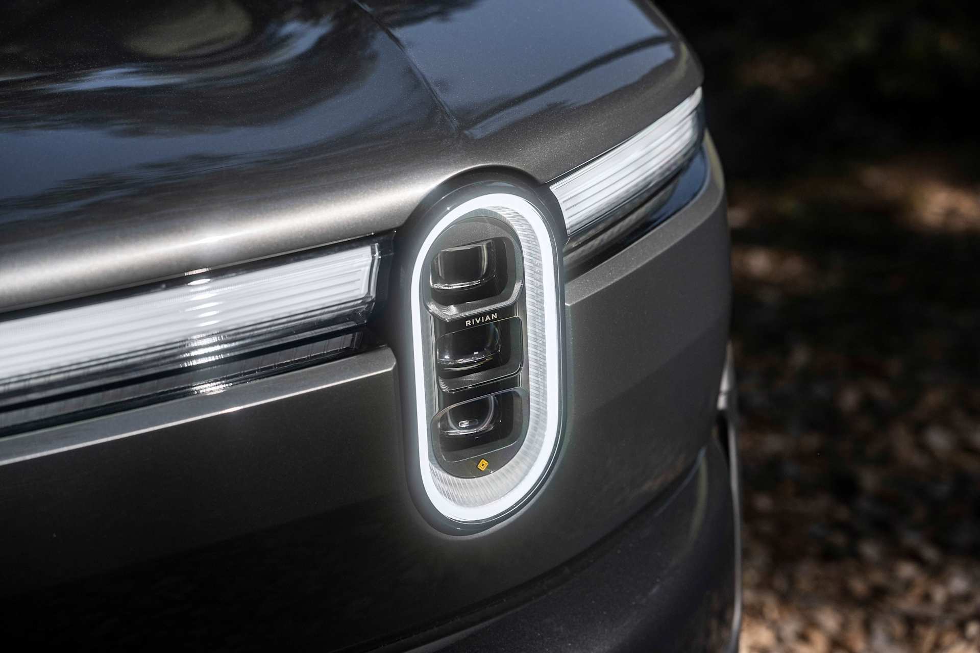 Close up of the front headlight of a gray Rivian truck. The light is outlined in a bright white LED strip. The name Rivian is printed on the light.