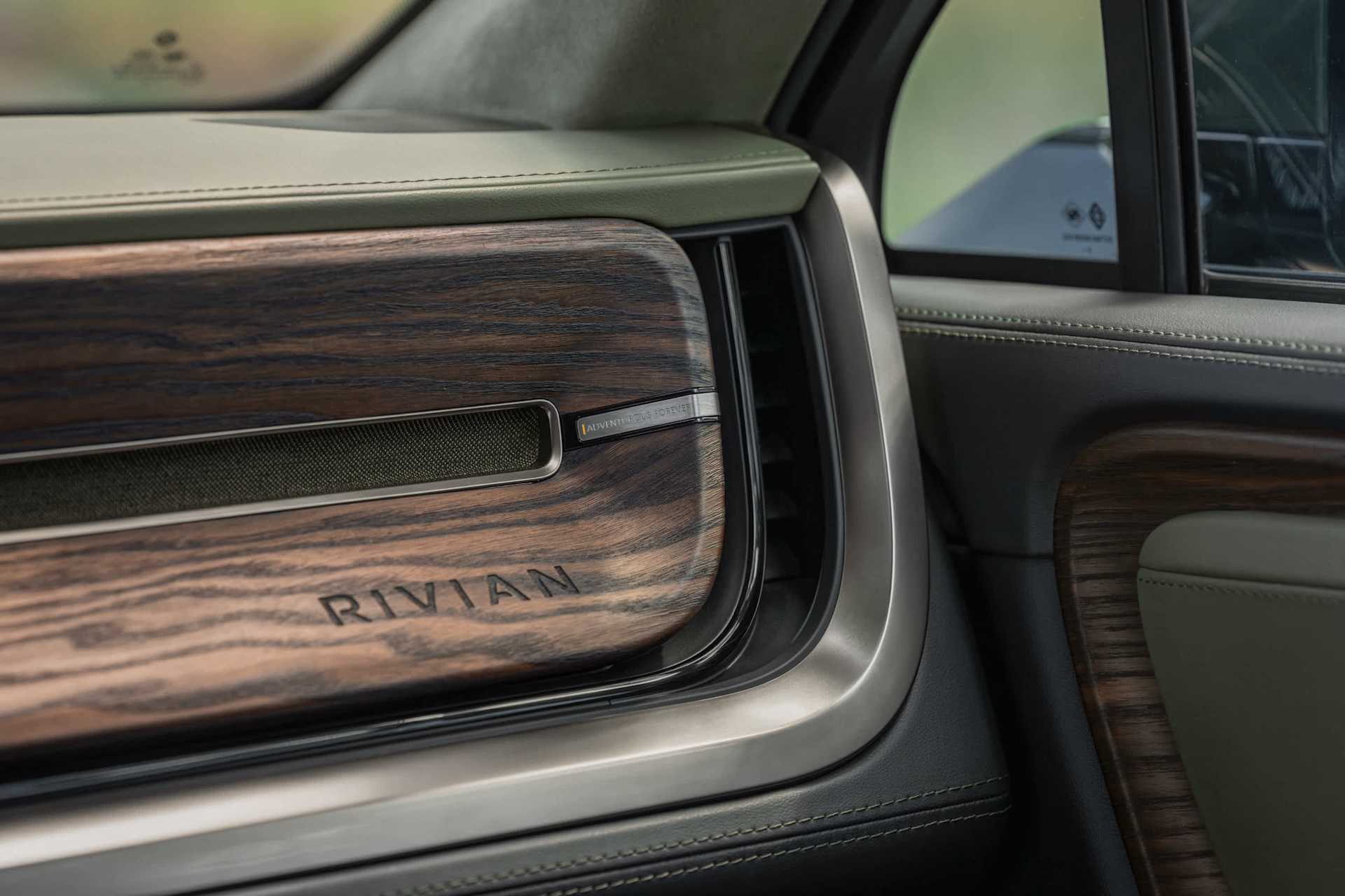 Rivian interior features dark wood grain paneling with the brand name etched on the surface. A speaker grill and "Adventure is Forever" detail are vis