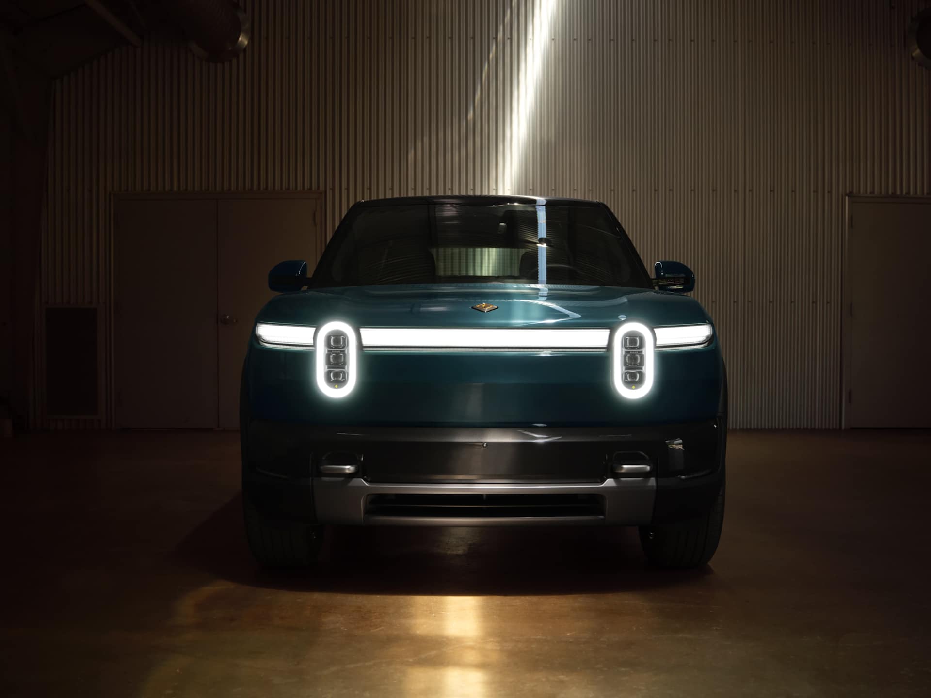 Front view of a teal Rivian R2 electric SUV in a garage with corrugated metal walls. The vehicle's headlights are illuminated, featuring a horizontal 