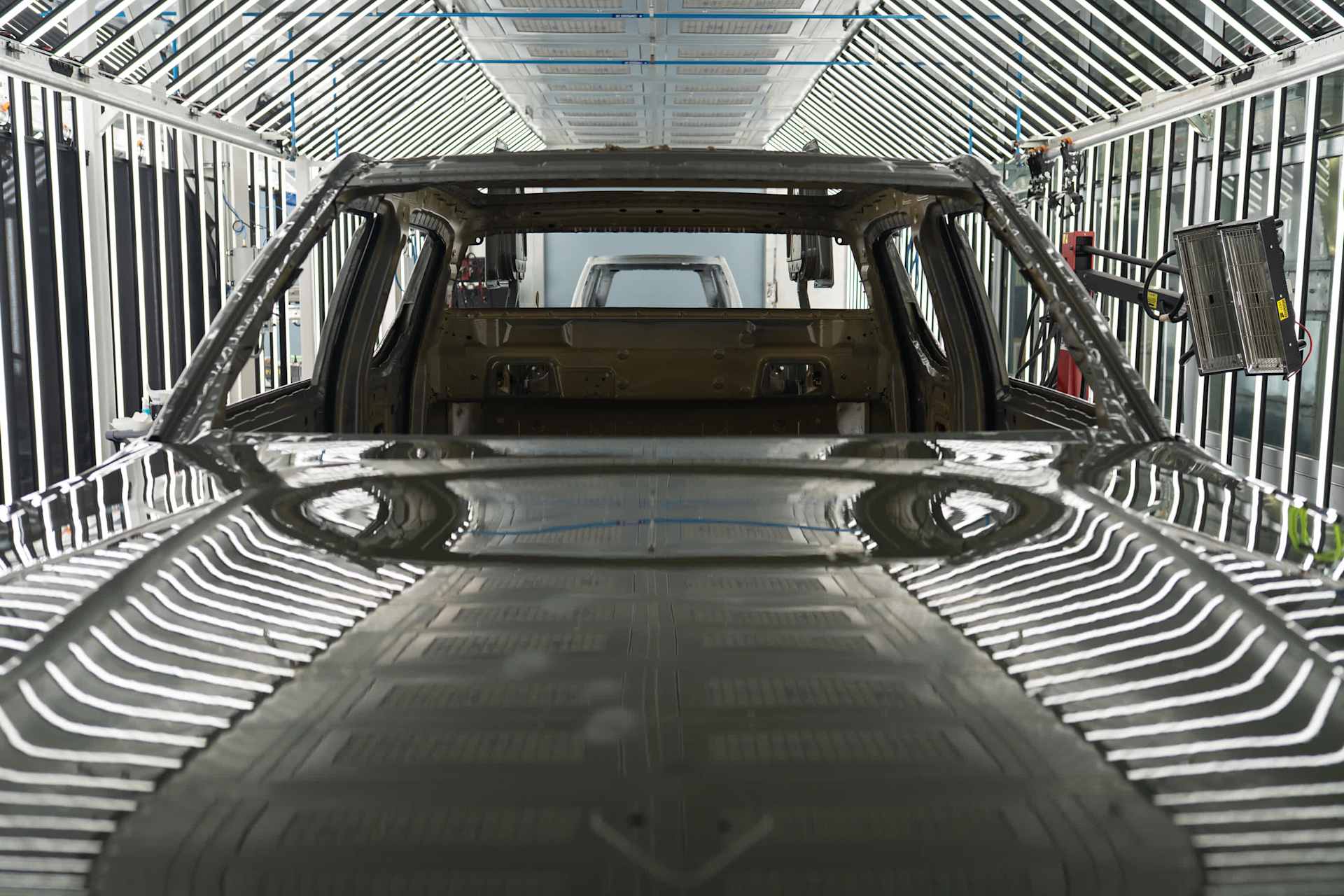 Rivian car frame moves through a factory assembly line. The vehicle is painted Rivian Green, reflecting overhead lights.
