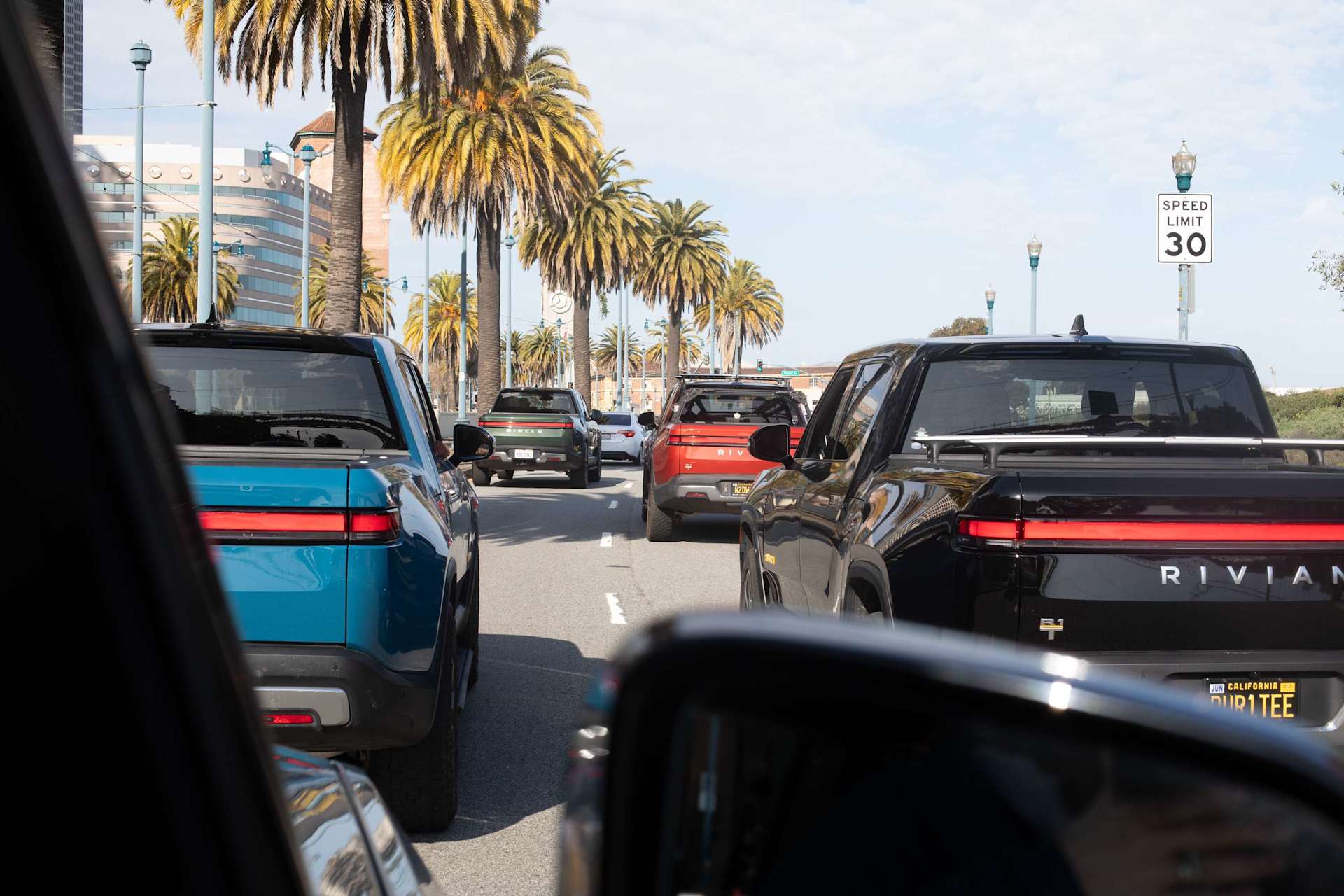 Rivian trucks in traffic on a sunny city street lined with palm trees. The trucks are blue, green, orange, and black. A speed limit sign reads "30".
