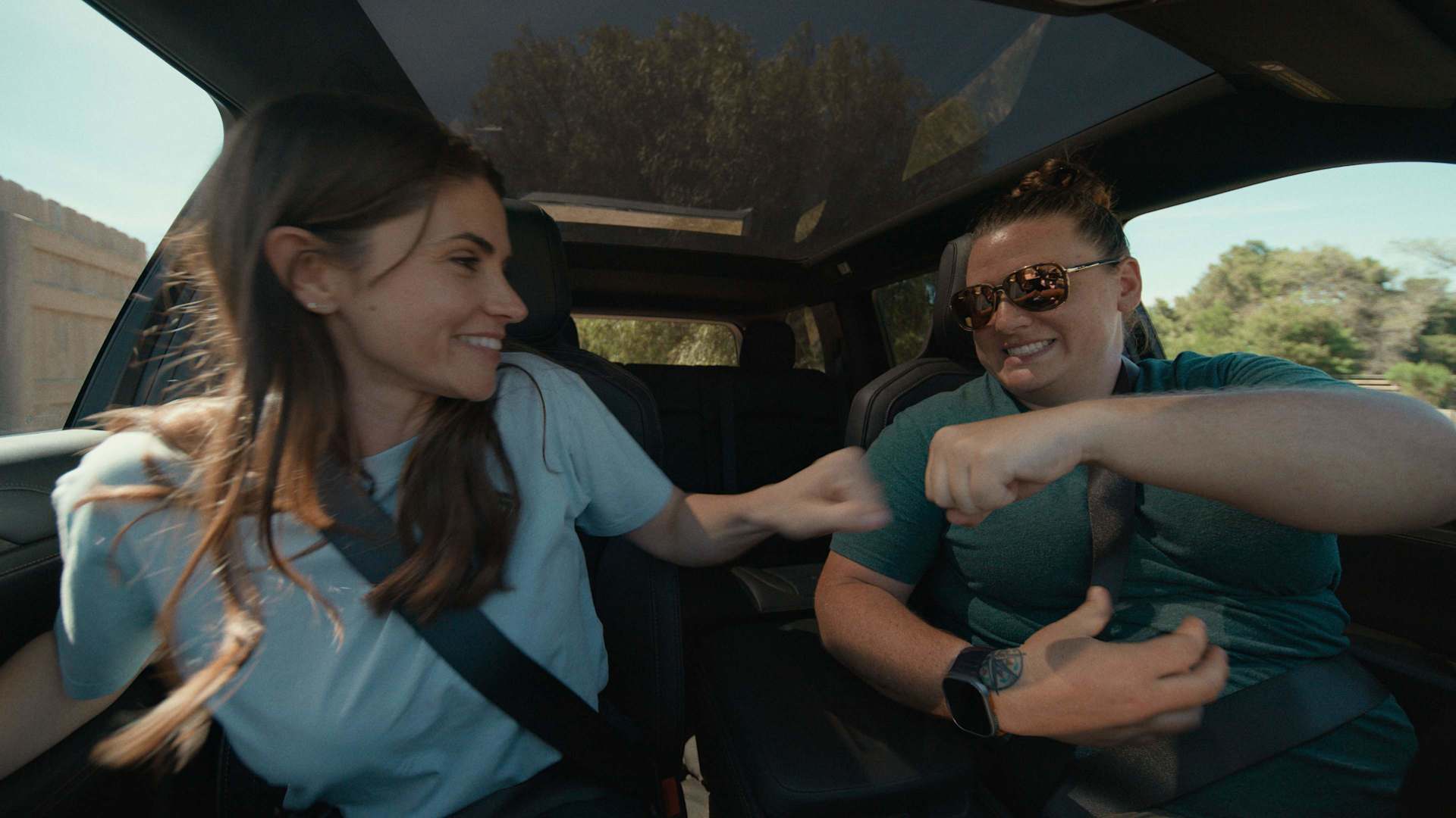 Two women fist-bump in a car with a glass roof. The woman on the left smiles, wearing a light blue shirt. The woman on the right wears sunglasses and 