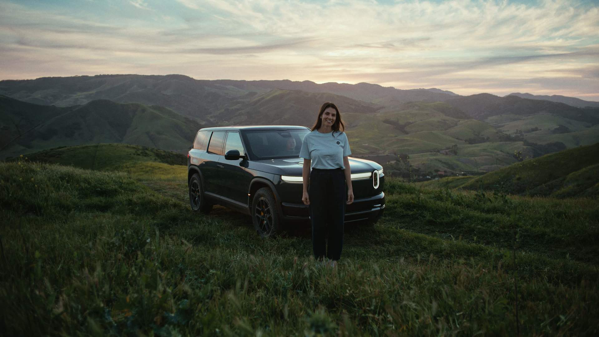 A woman stands smiling in front of a Rivian SUV on a grassy hillside. Rolling green hills stretch into the distance under a cloudy sky at dusk.