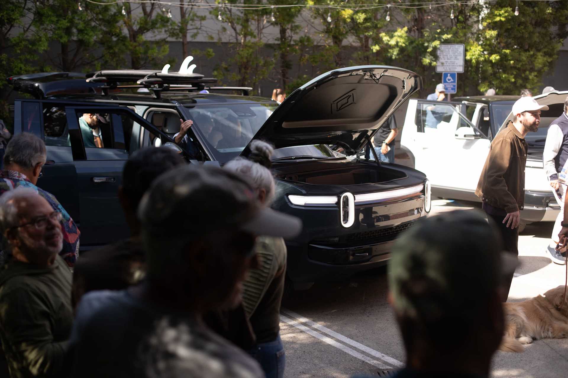 Dark green Rivian SUV with hood open and surfboards on top is surrounded by a crowd of people. The front lights of the car are on. The vehicle is park