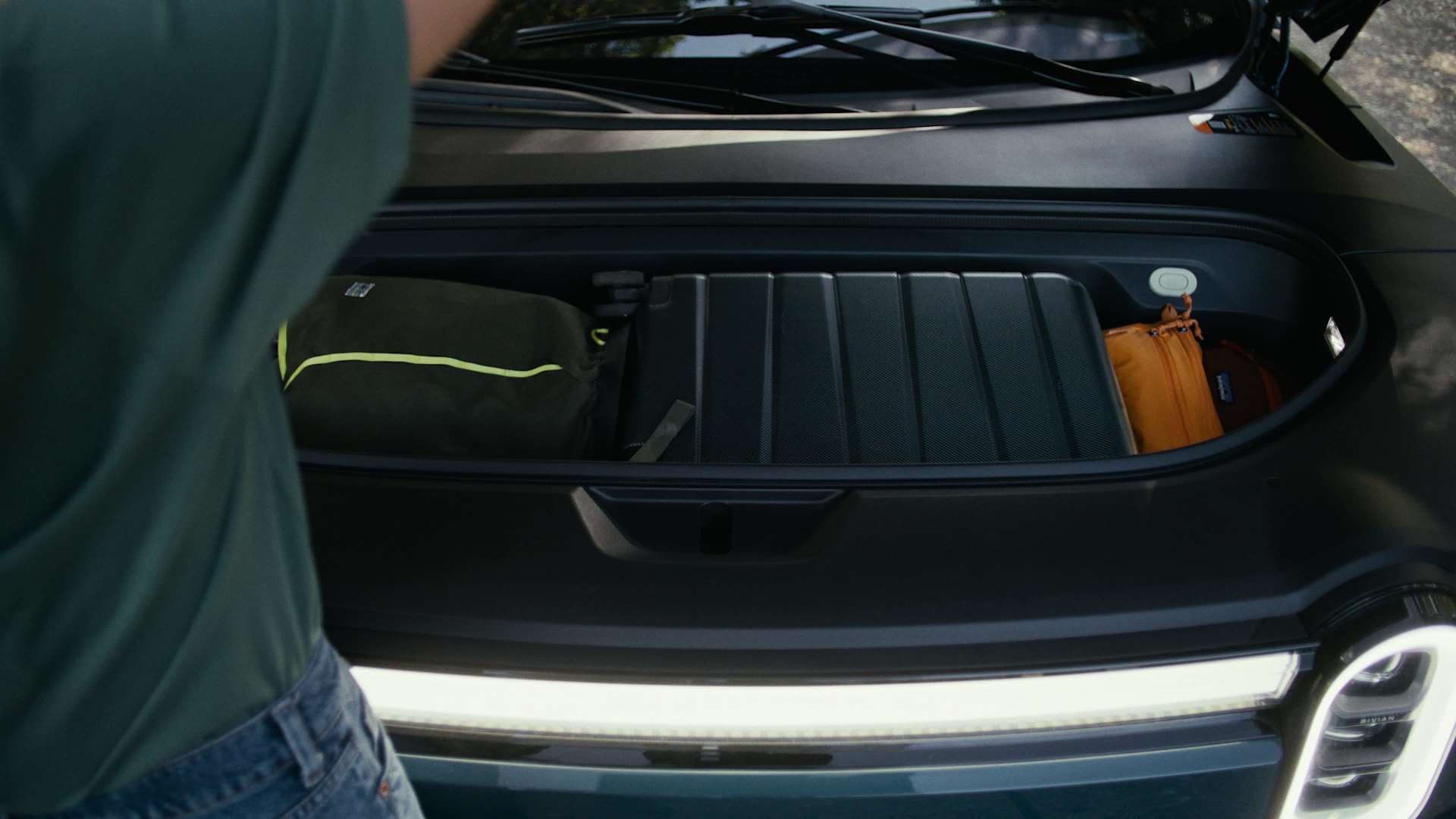 Rivian R1T's front trunk, or "frunk," is open and filled with luggage. A black suitcase sits in the center, with a green duffel bag on the left and an