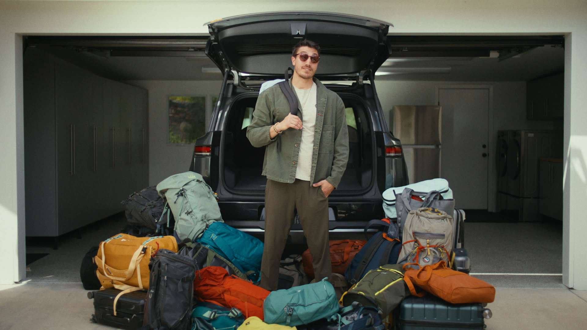 Man stands in garage with open SUV trunk behind a pile of luggage. He wears sunglasses, a green jacket, and has a backpack slung over his shoulder. Ne