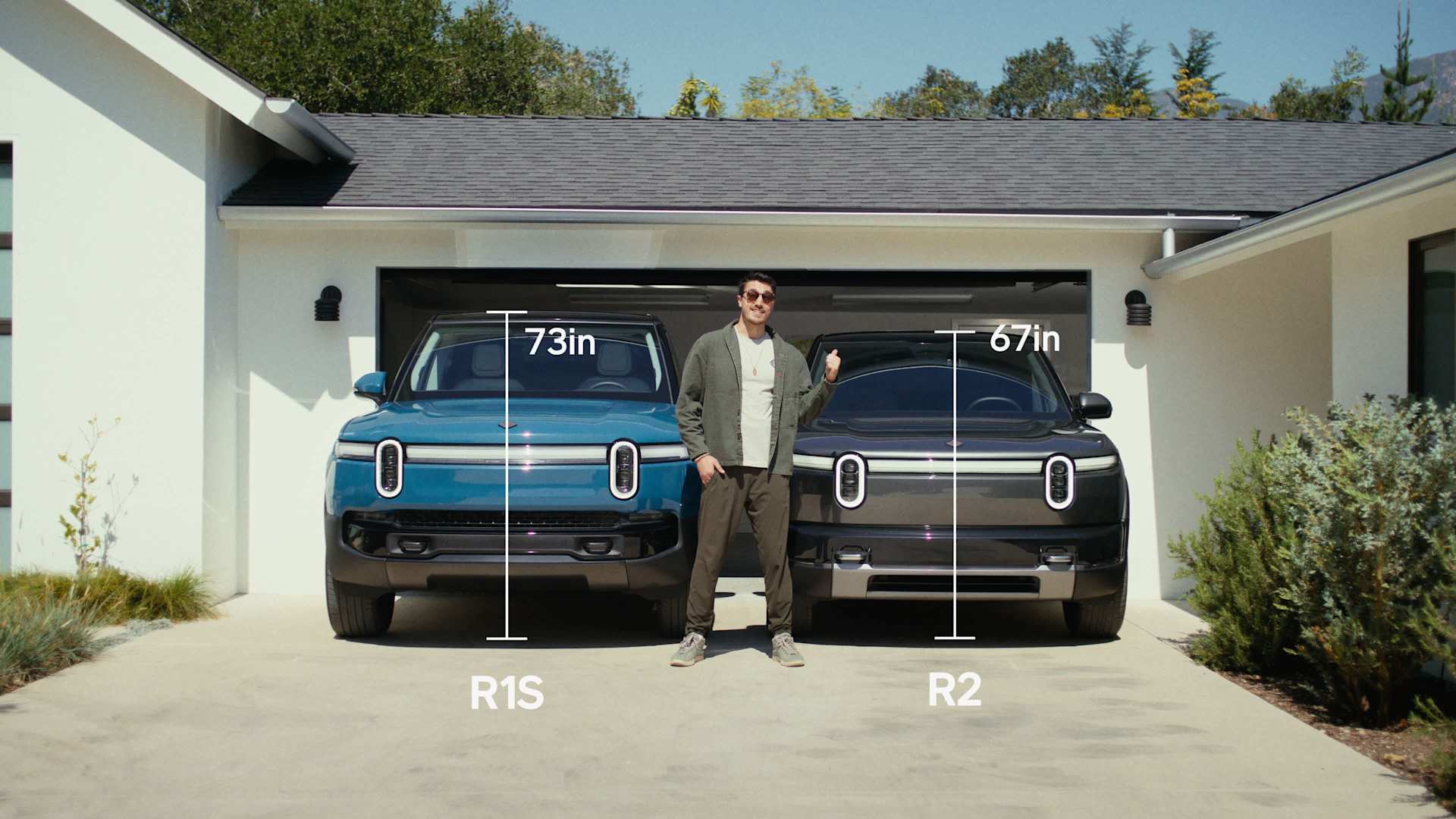 A man stands between a blue R1S (73in) and a gray R2 (67in) in a garage. The man points at the R2, indicating its height. The garage is white with a d