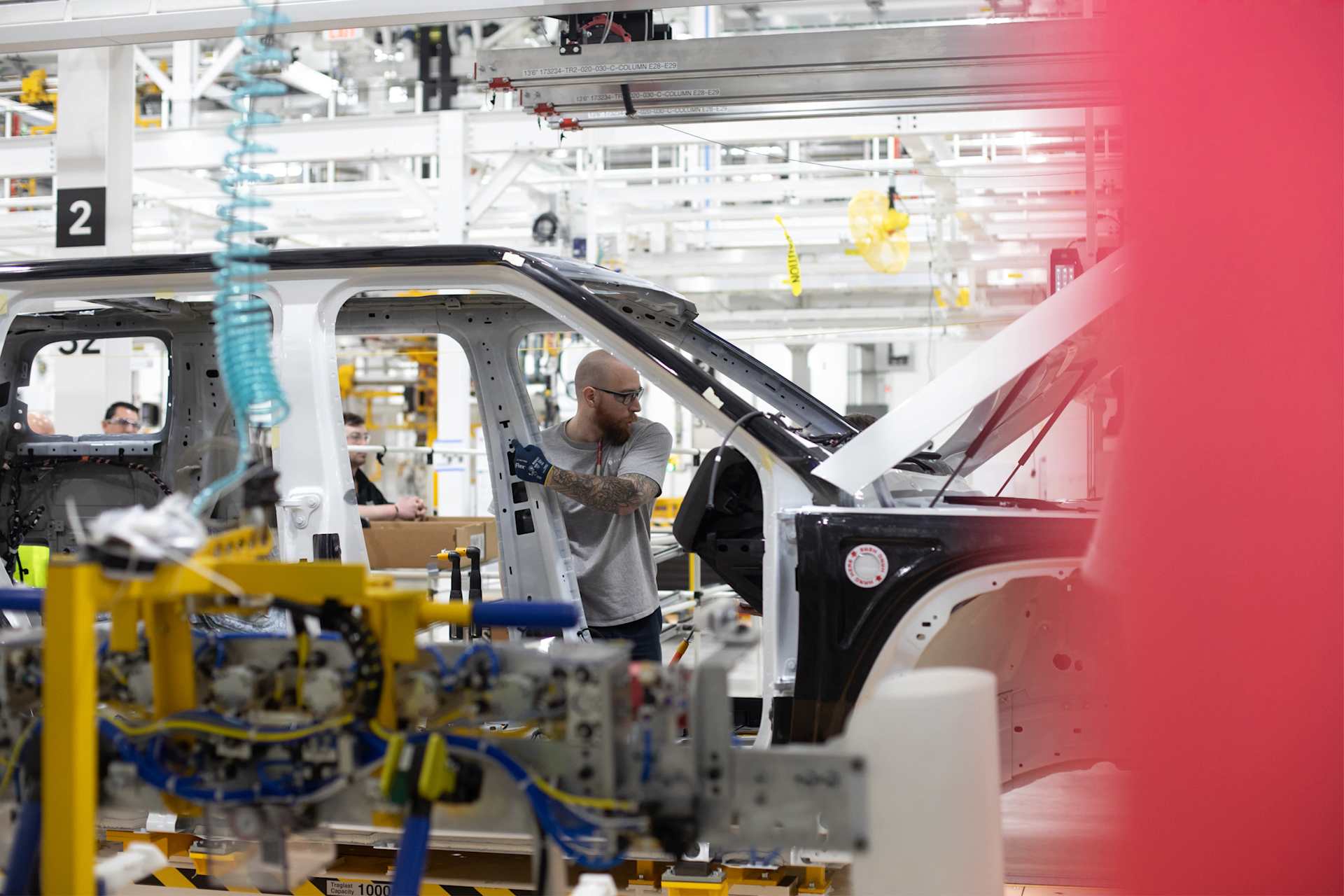 A factory worker in a gray t-shirt and glasses works on the frame of a white car. The car frame is on an assembly line in a bright factory. The factor