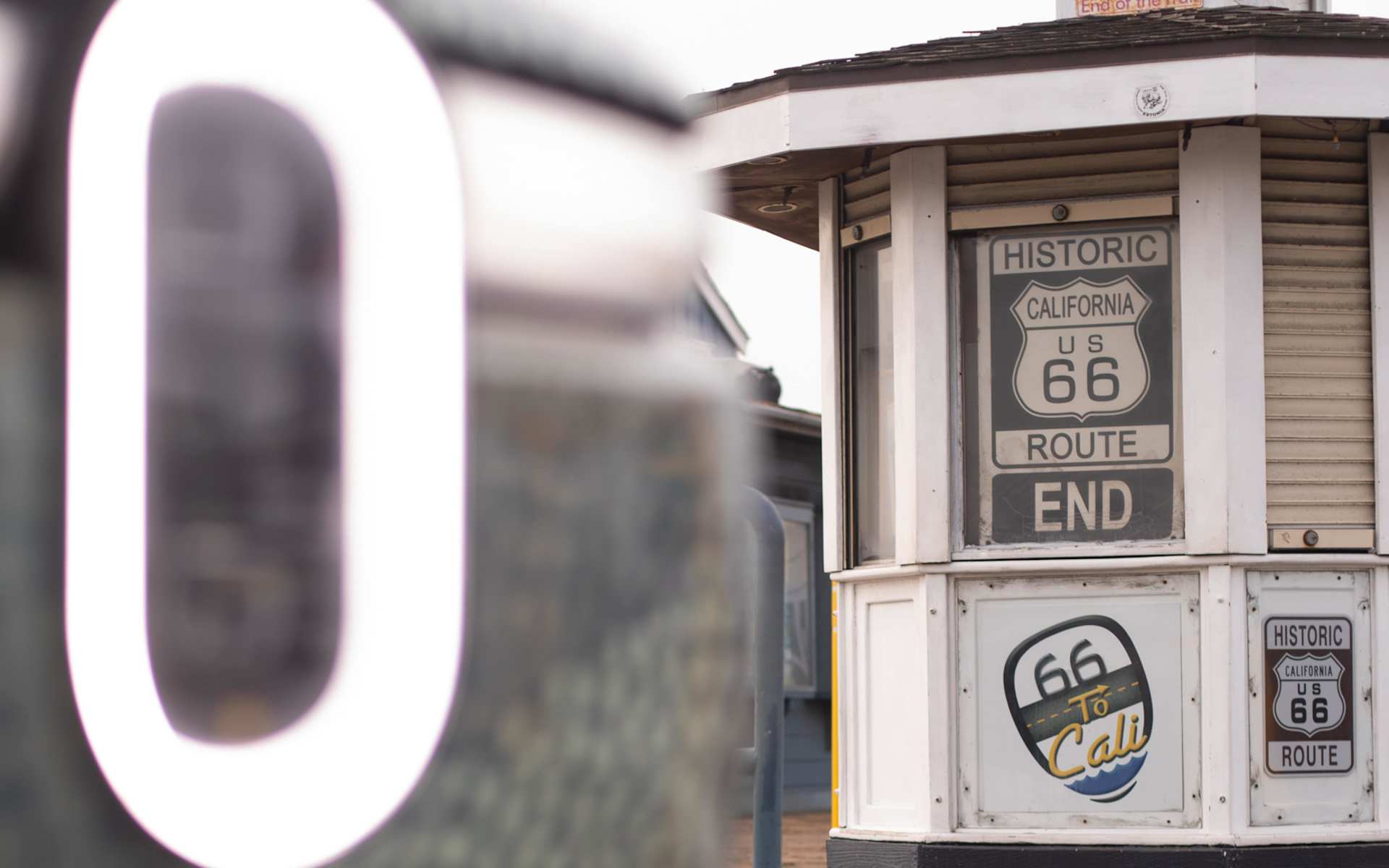A white Route 66 end of the road kiosk with "Historic US Route 66 End" sign. A blurred, illuminated "O" is in the foreground.